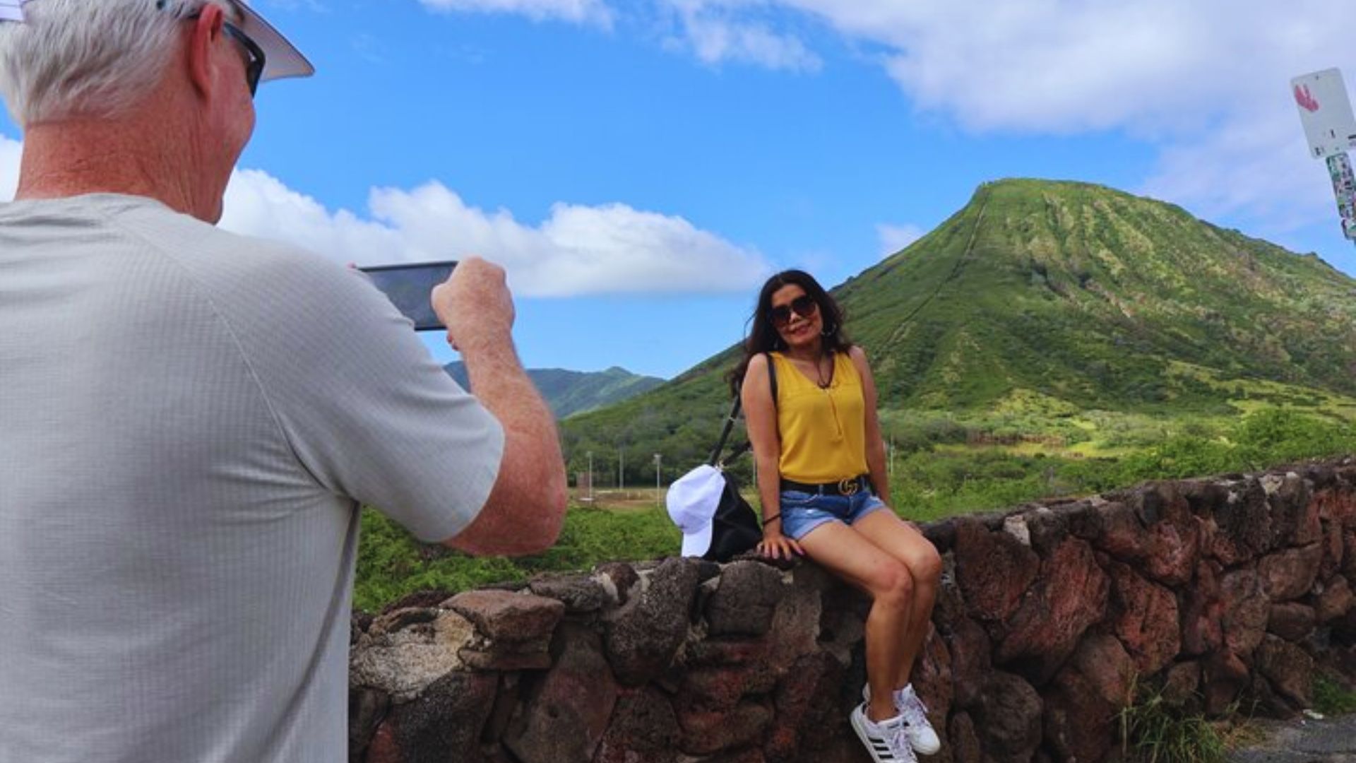 A man photographs a woman sitting casually on a stone wall in a sunny outdoor setting.
