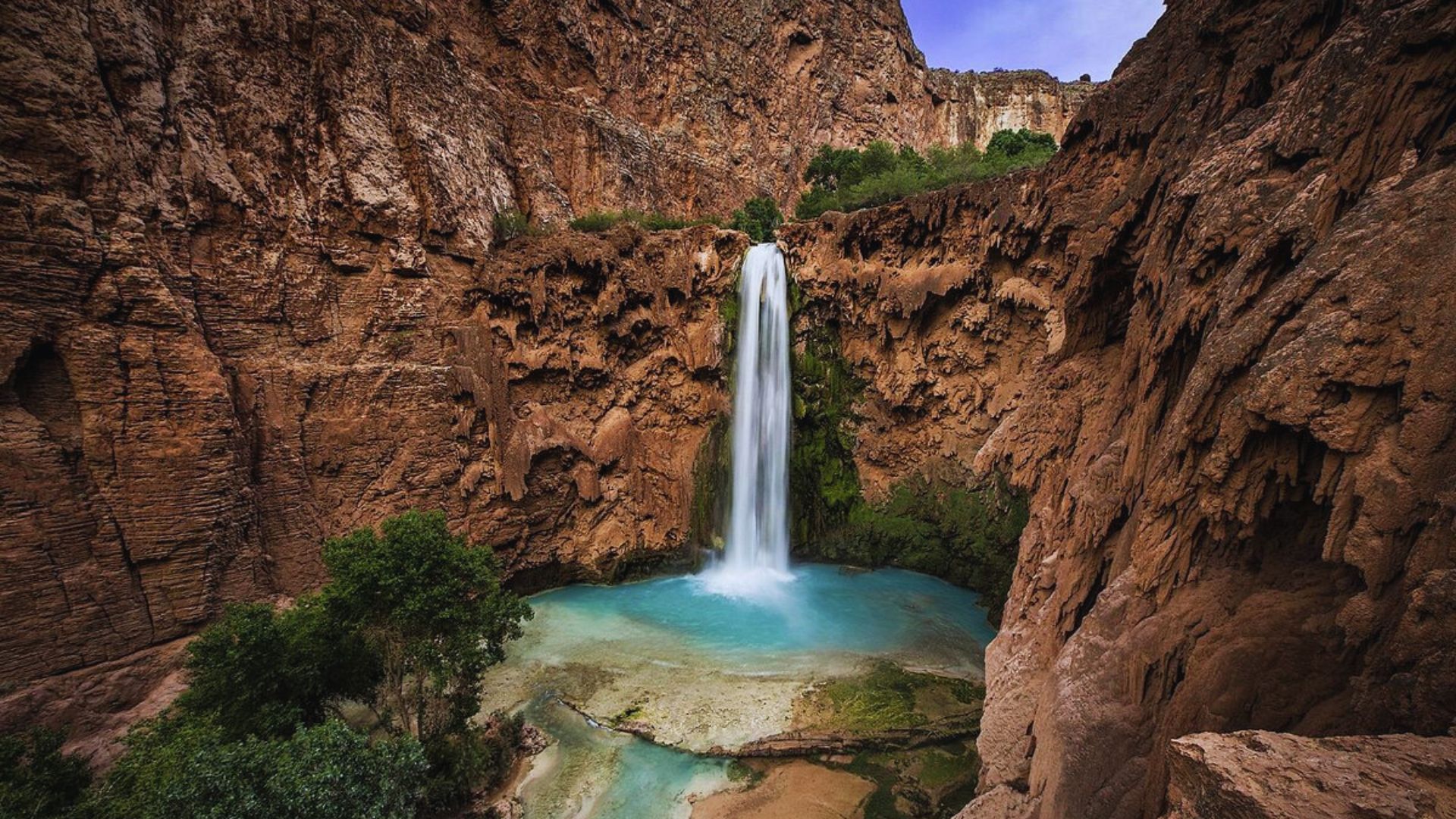 A stunning waterfall cascades into a turquoise pool surrounded by rugged red cliffs and lush greenery under a clear blue sky, creating a serene and majestic scene.