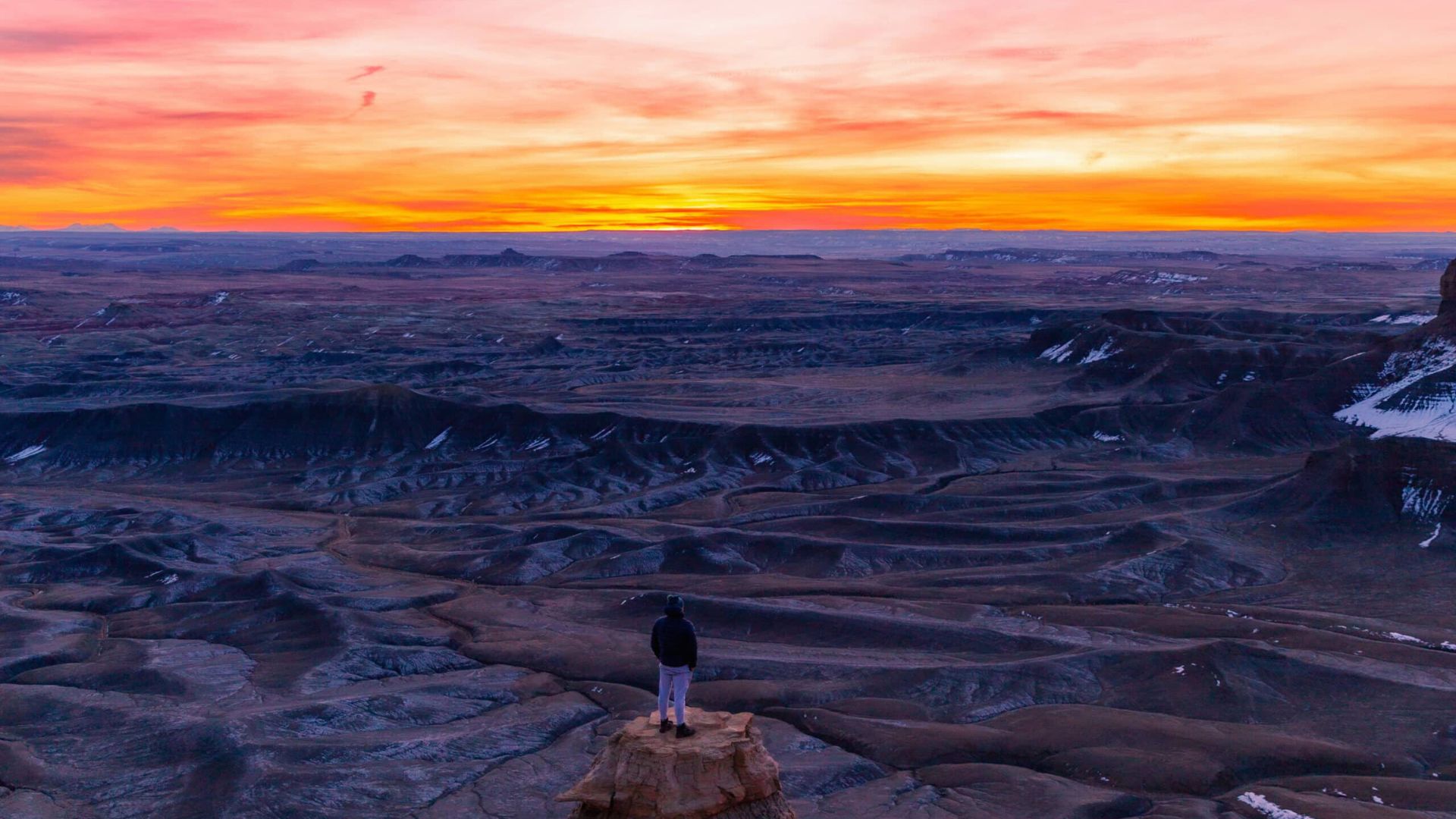 A person stands on a rock, overlooking a vast, rugged desert landscape at sunset. The sky is ablaze with orange and pink hues, conveying serenity.