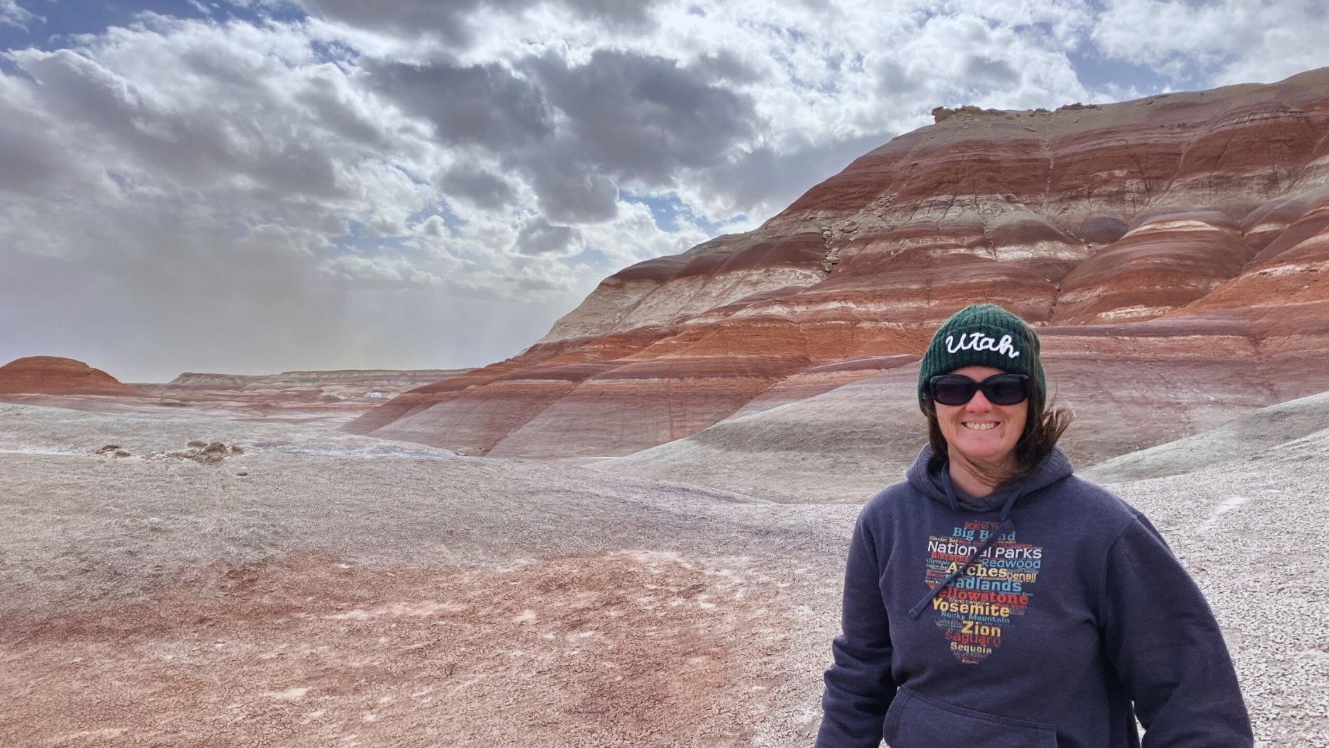 A person wearing sunglasses and a beanie with "Utah" text stands smiling in front of colorful, layered rocky hills under a cloudy sky.