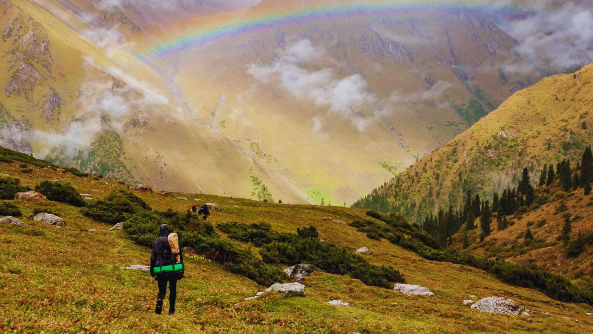 A person ascends a mountain trail beneath a vibrant rainbow arching across the sky.