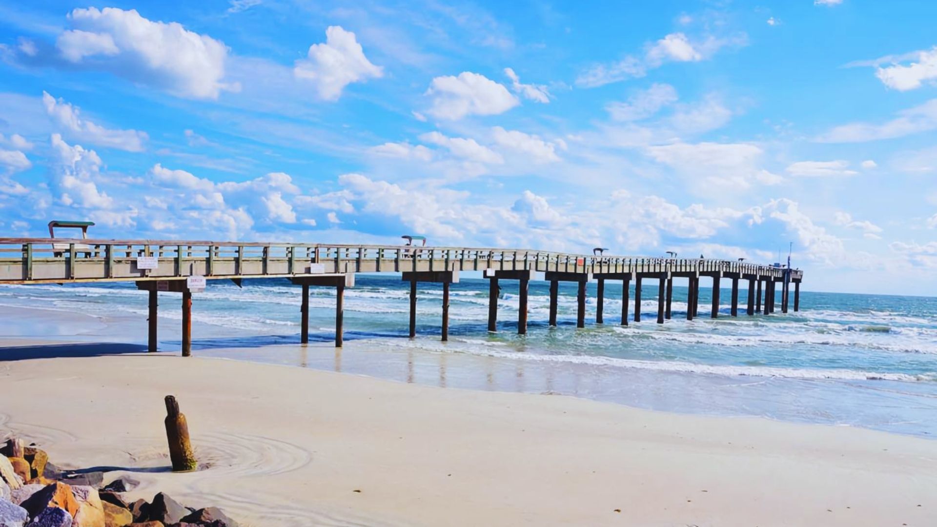 A scenic pier on the beach with a vibrant blue sky and soft white clouds above.
