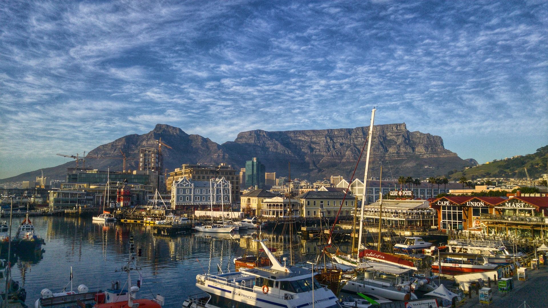 A scenic harbor with boats docked in front of a majestic mountain backdrop under a clear blue sky.