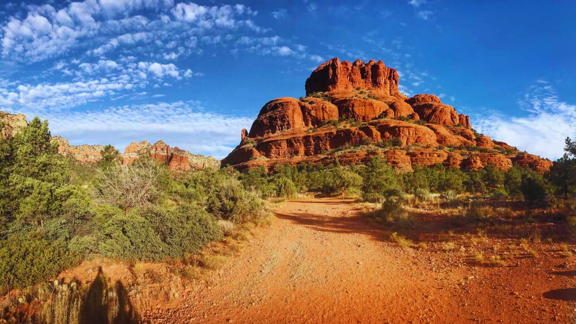 A scenic view of Bell Rock in Sedona, Arizona, under a bright blue sky with wispy clouds. The red rock formation is surrounded by green shrubs.