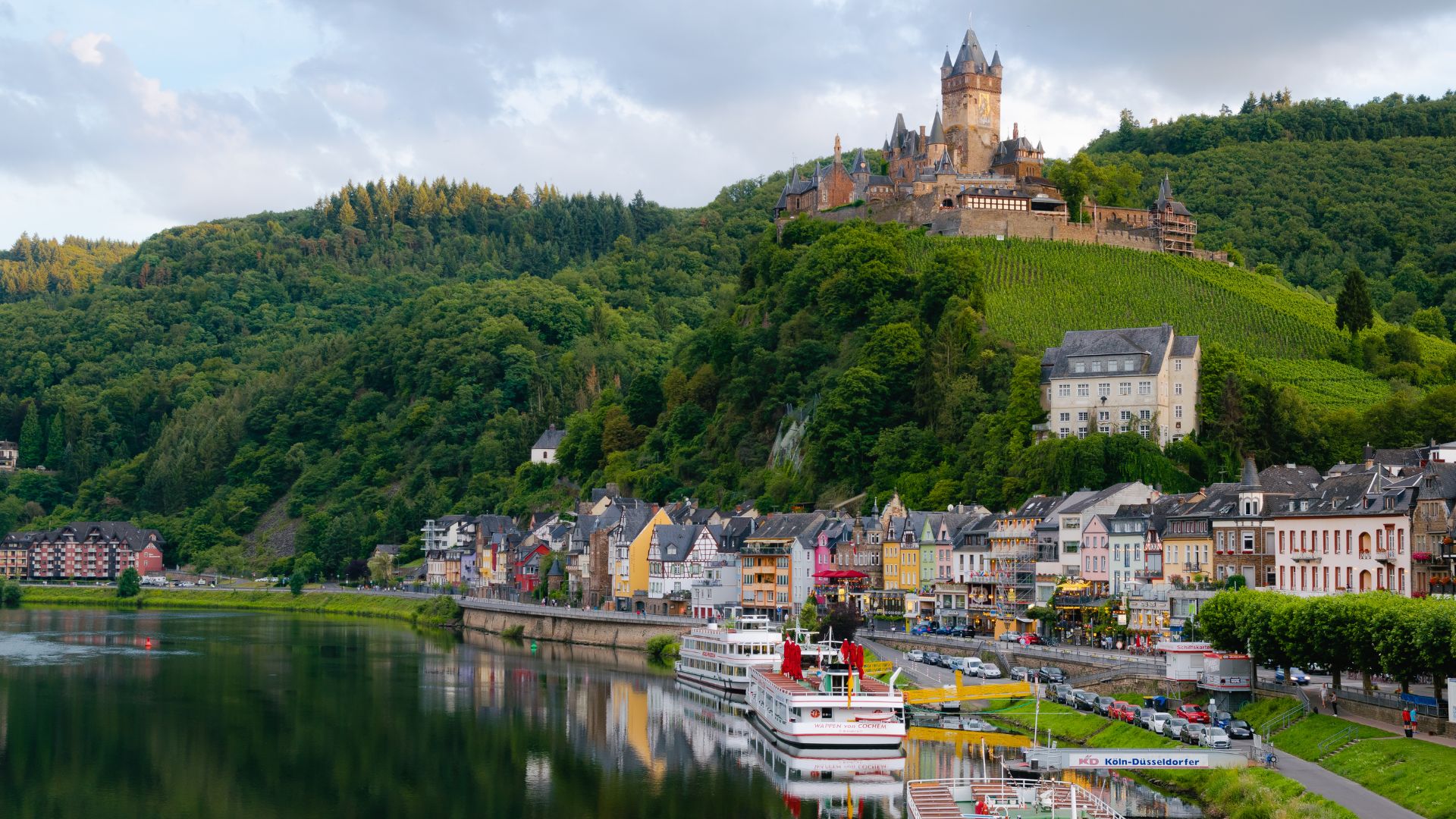A picturesque view of a riverfront town with colorful buildings next to the water. A grand castle sits atop a lush green hill under a partly cloudy sky.