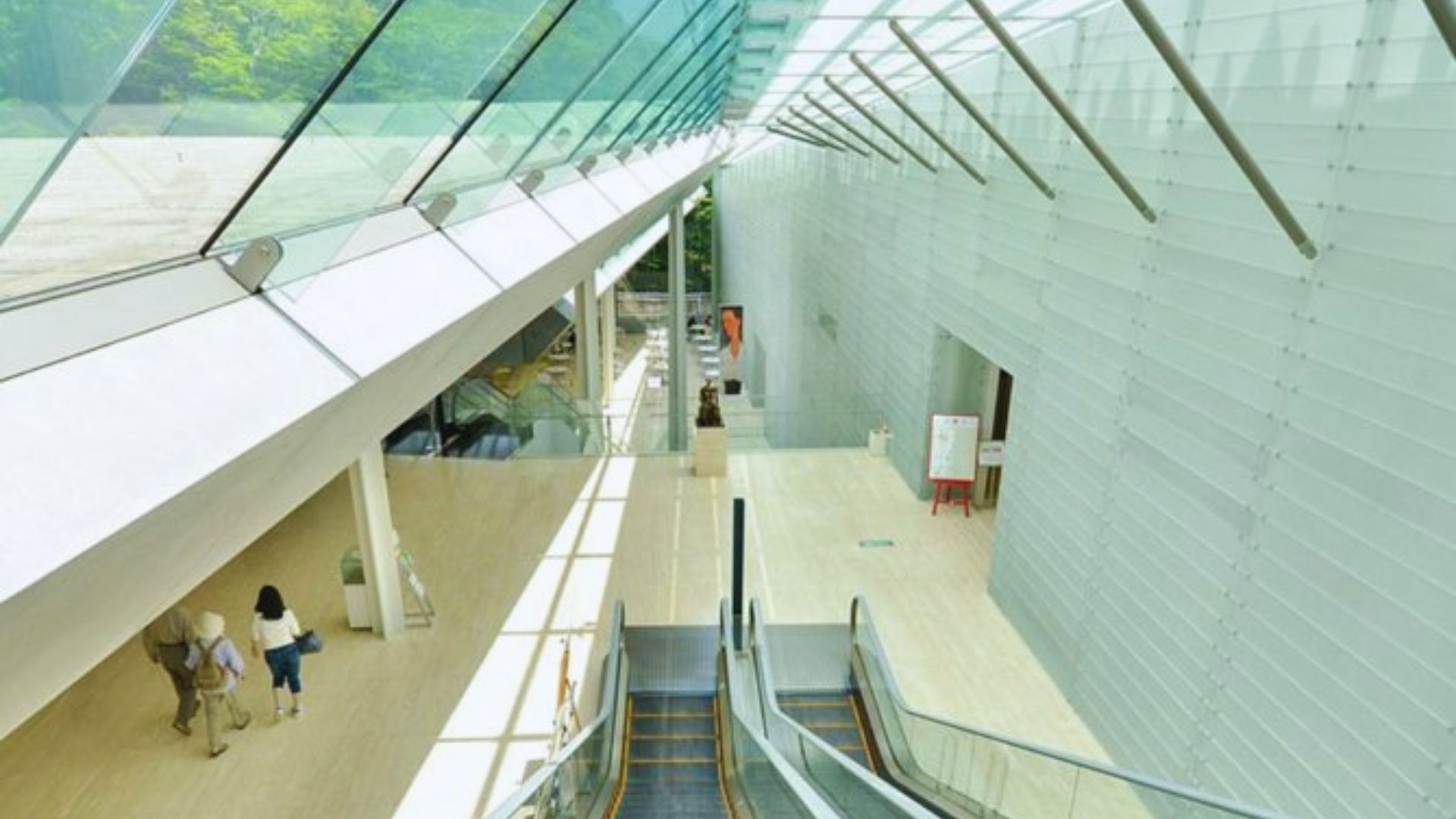 Modern interior with escalators leading down a bright, spacious corridor. Glass ceiling and white walls, with people walking on the left. Peaceful ambiance.