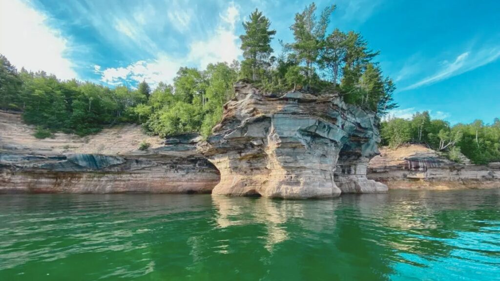 Rocky cliffs with layered formations rise from a green lake, topped with lush trees under a vivid blue sky, conveying a serene and majestic natural landscape.