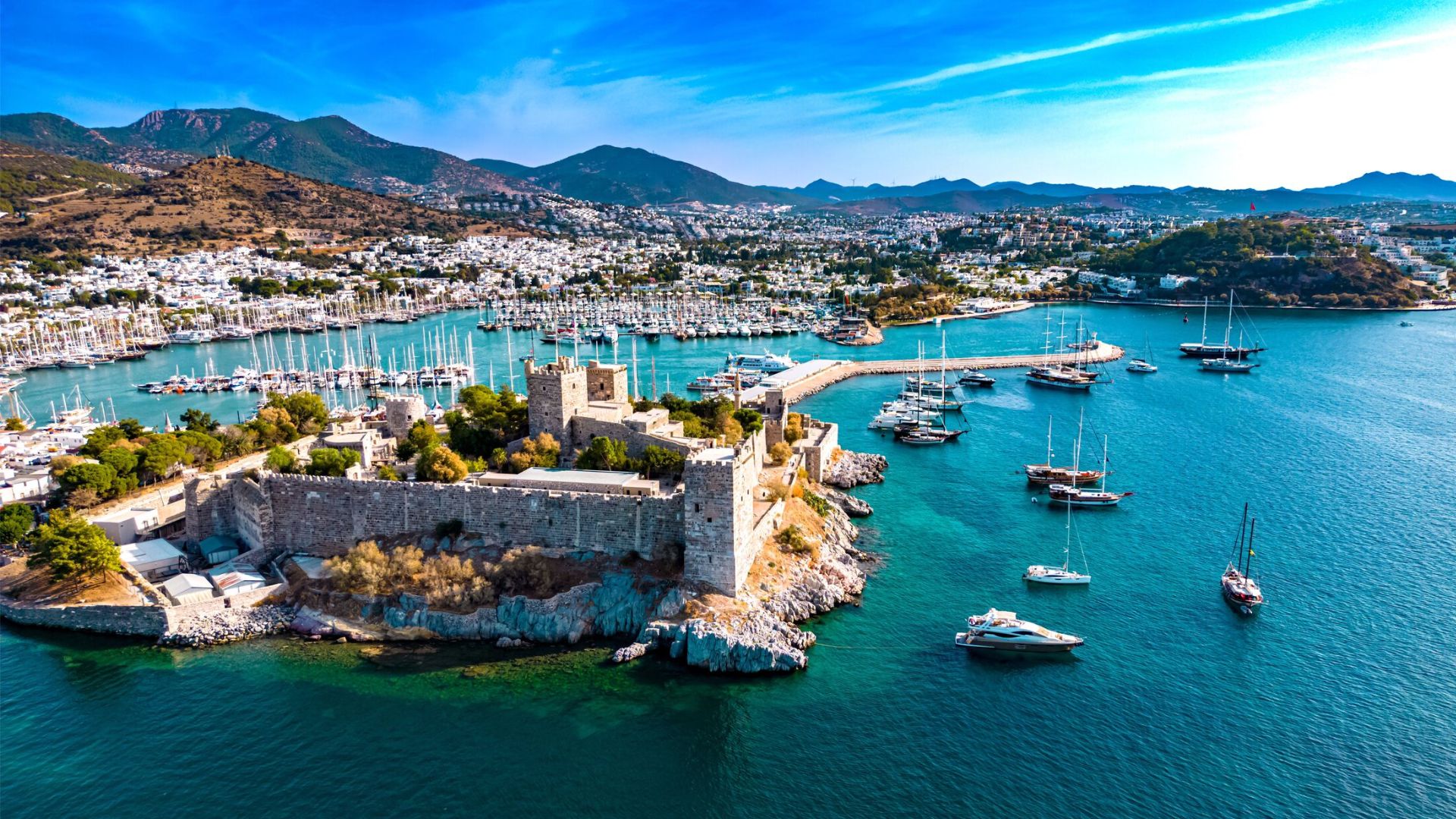 Aerial view of a coastal town featuring a historic stone castle surrounded by clear blue water, sailboats, and a backdrop of hills under a vibrant sky.