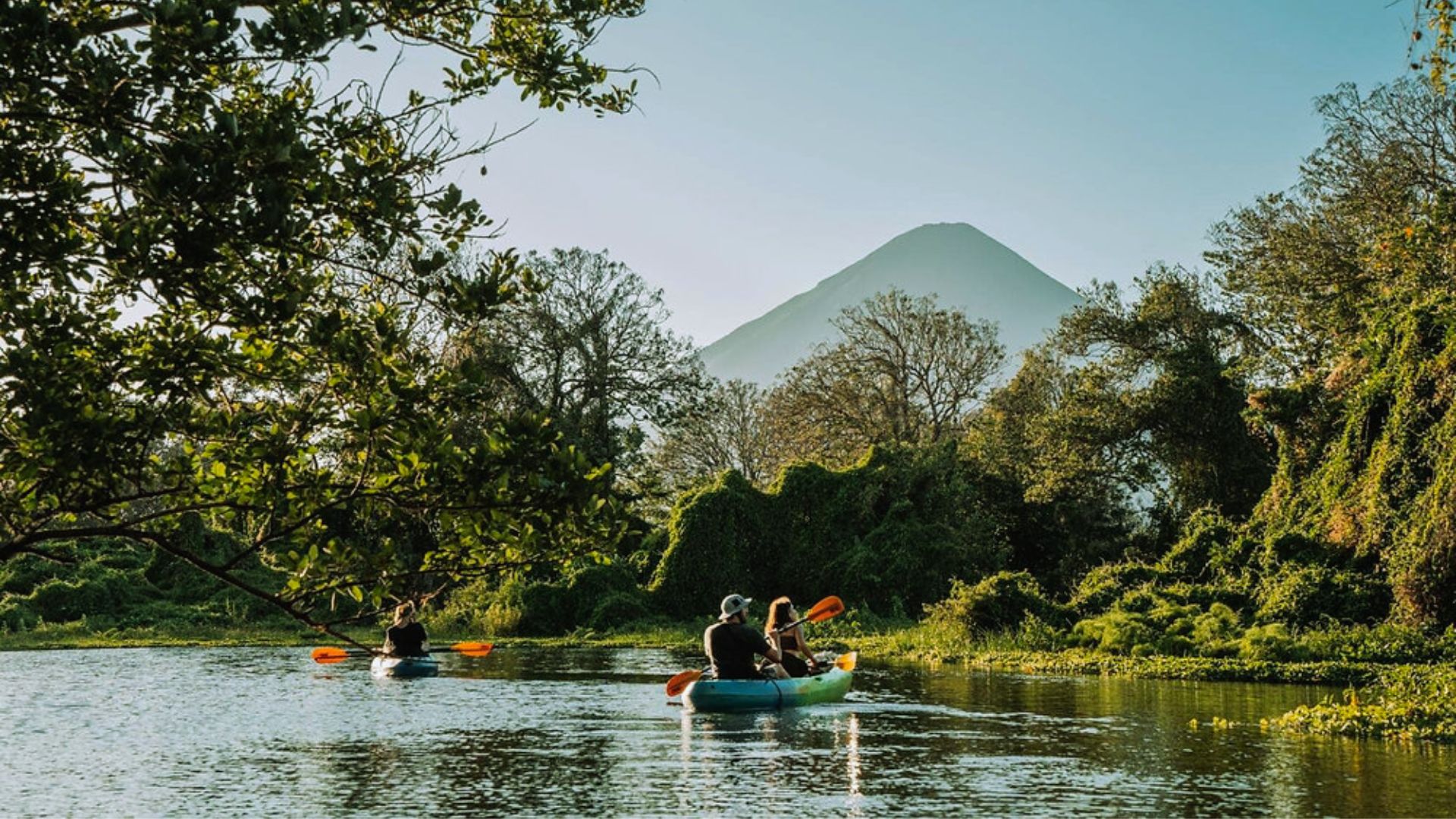 Two people kayaking on a serene river surrounded by lush greenery and trees, with a distant volcano under a clear blue sky, conveying tranquility.