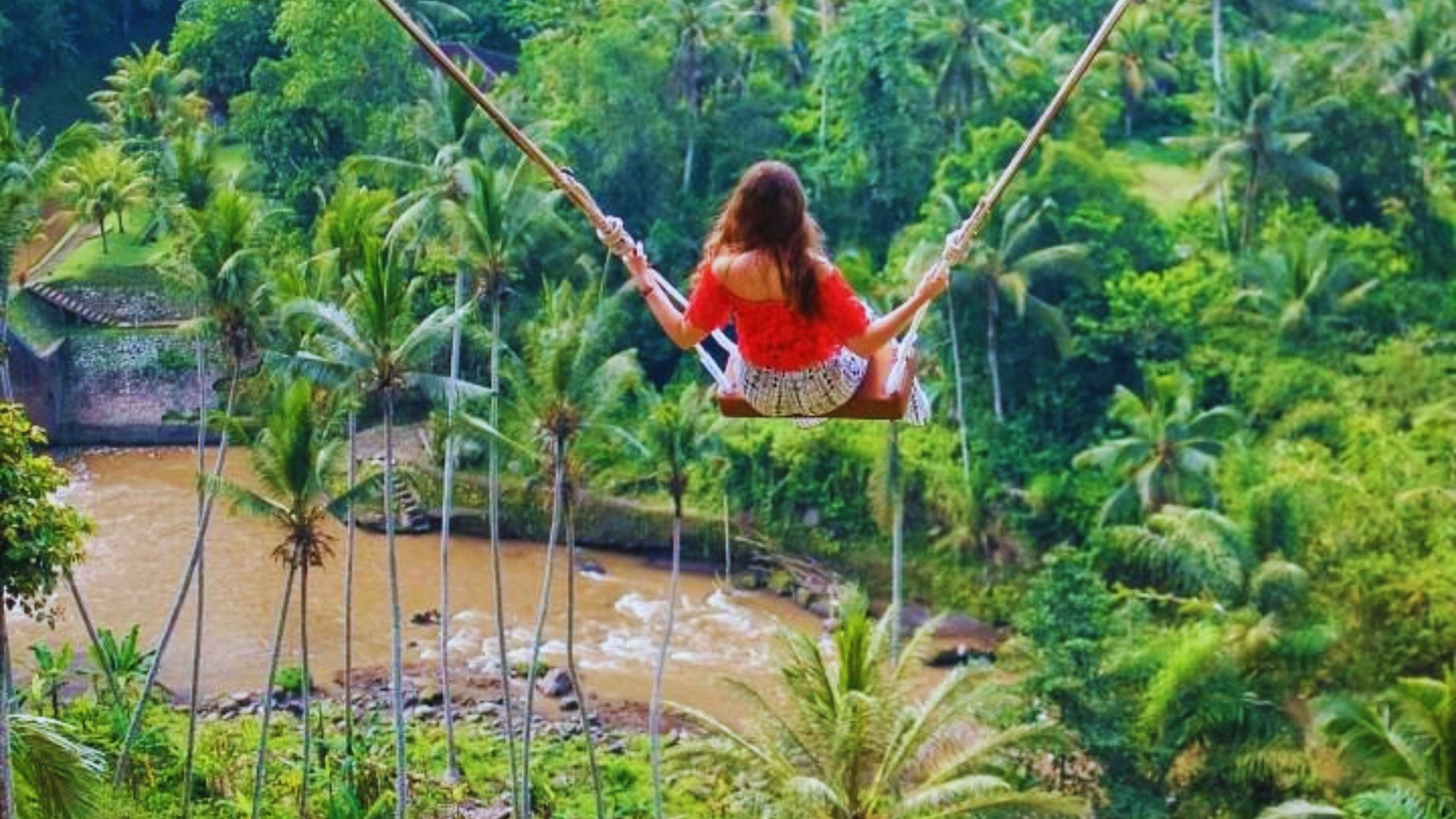 A woman in a red top and patterned skirt is on a swing high above a lush green jungle with palm trees and a winding brown river below, evoking a sense of adventure.
