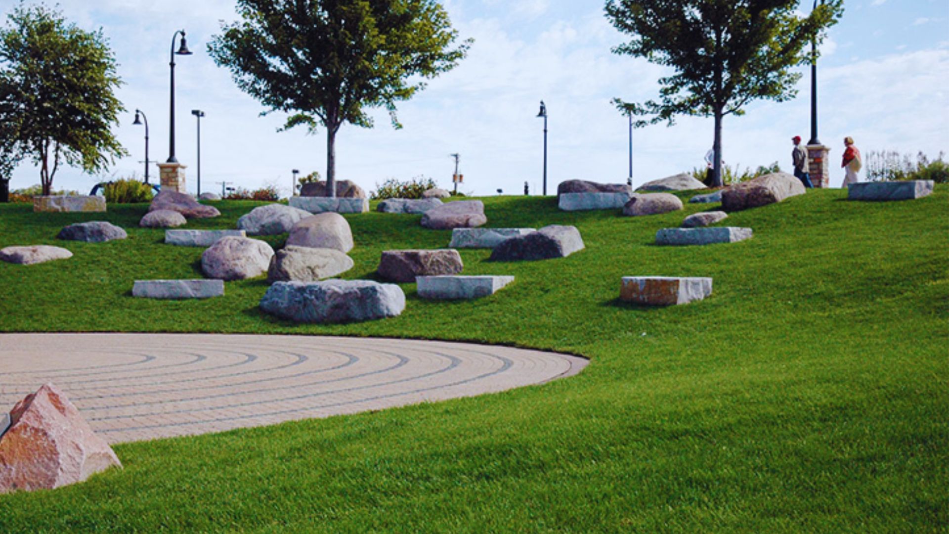 A peaceful park scene with scattered large rocks on a grassy hill under a blue sky. Two people are walking near trees in the background.