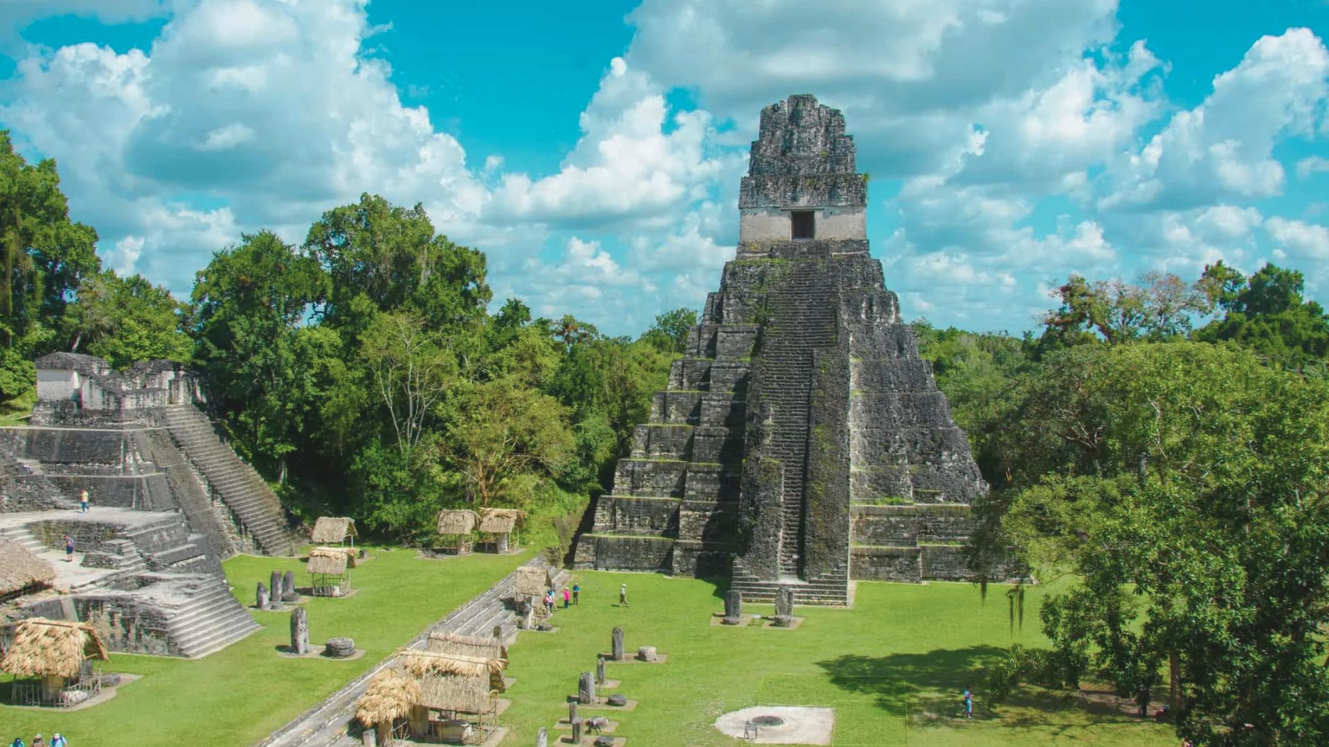 Ancient Mayan pyramid surrounded by lush jungle and scattered ruins under a bright blue sky with fluffy clouds, evoking a sense of mystery and history.