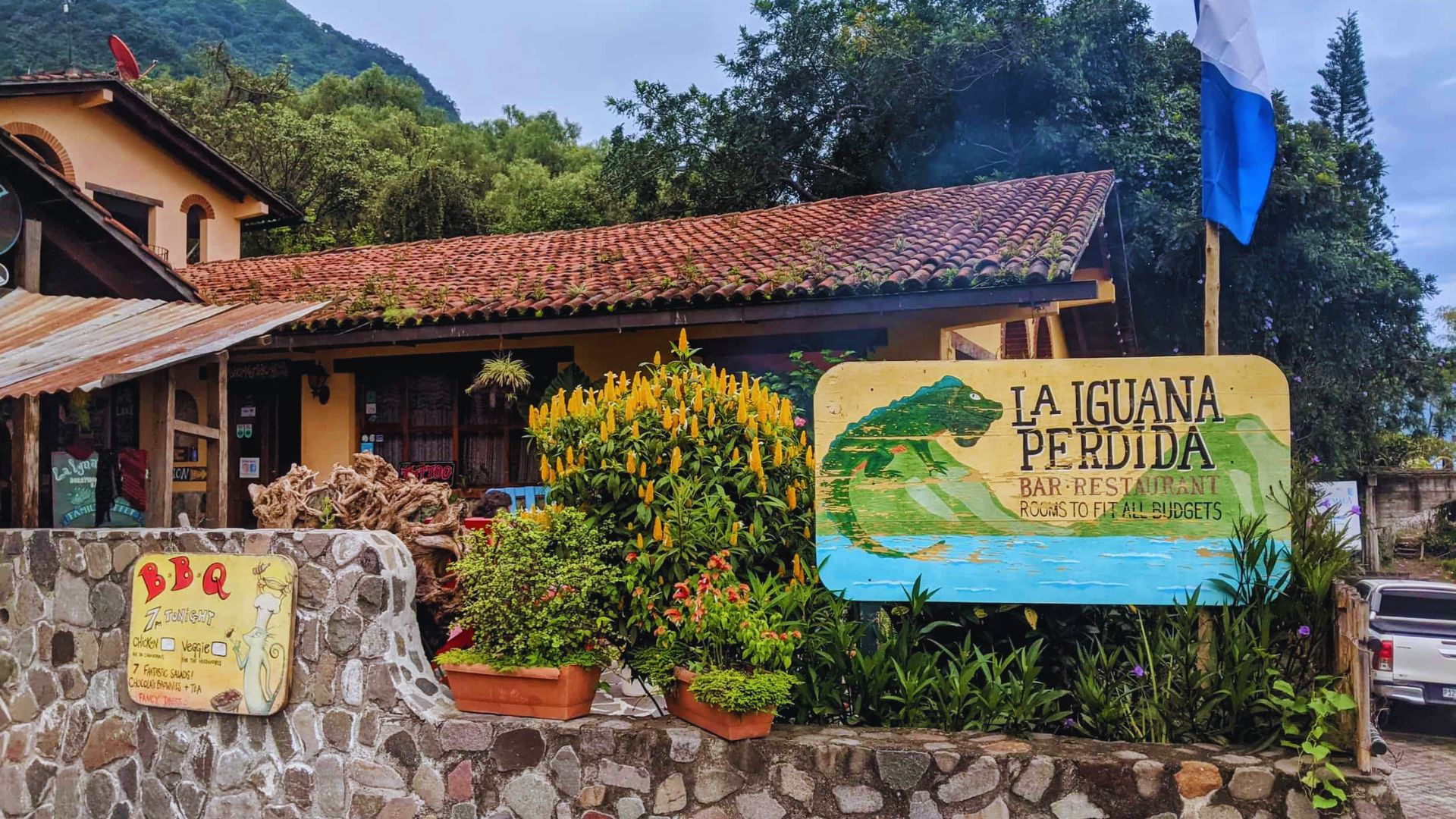 Rustic restaurant "La Iguana Perdida" with a tiled roof, nestled in lush greenery. A colorful sign and bright flowers create a welcoming ambiance.