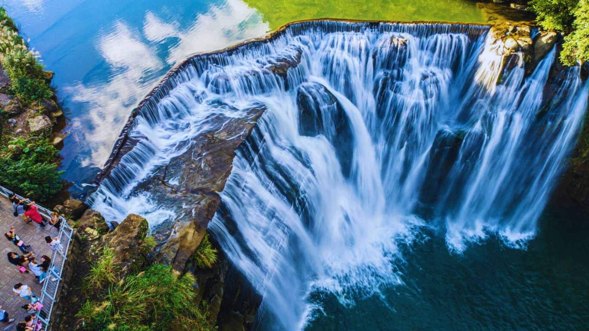Aerial view of a majestic waterfall cascading into a turquoise pool. Visitors stand on a stone platform, enjoying the view. The scene is vibrant and dynamic.