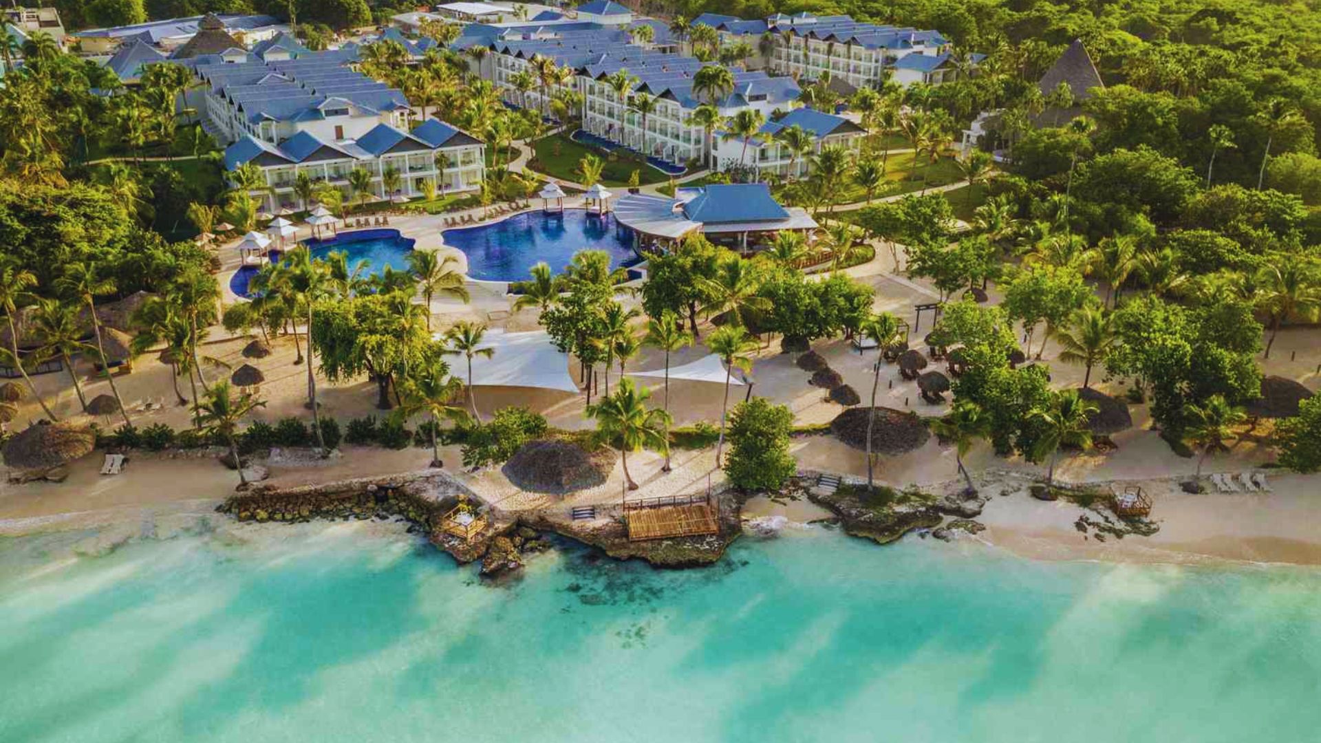 Aerial view of a tropical resort with blue-roofed buildings, a central pool, lush greenery, and a sandy beach next to turquoise ocean waves.