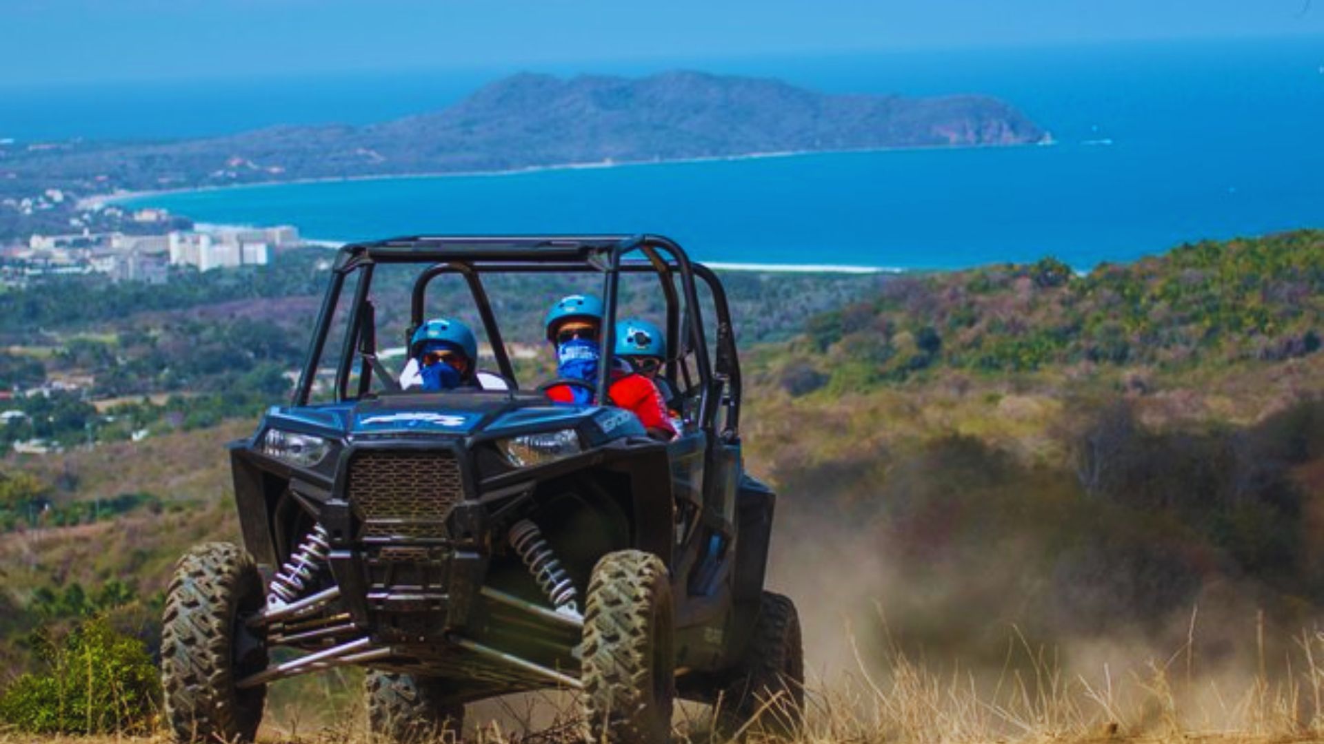 Two people in helmets ride a rugged off-road vehicle on a dusty trail overlooking a scenic coastal landscape, conveying adventure and excitement.