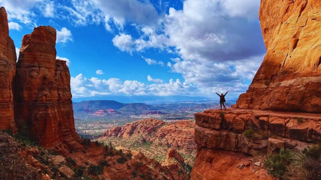 A person stands triumphantly on a red rock cliff in Sedona, Arizona. Vast desert landscape and dramatic clouds stretch under a vibrant blue sky.