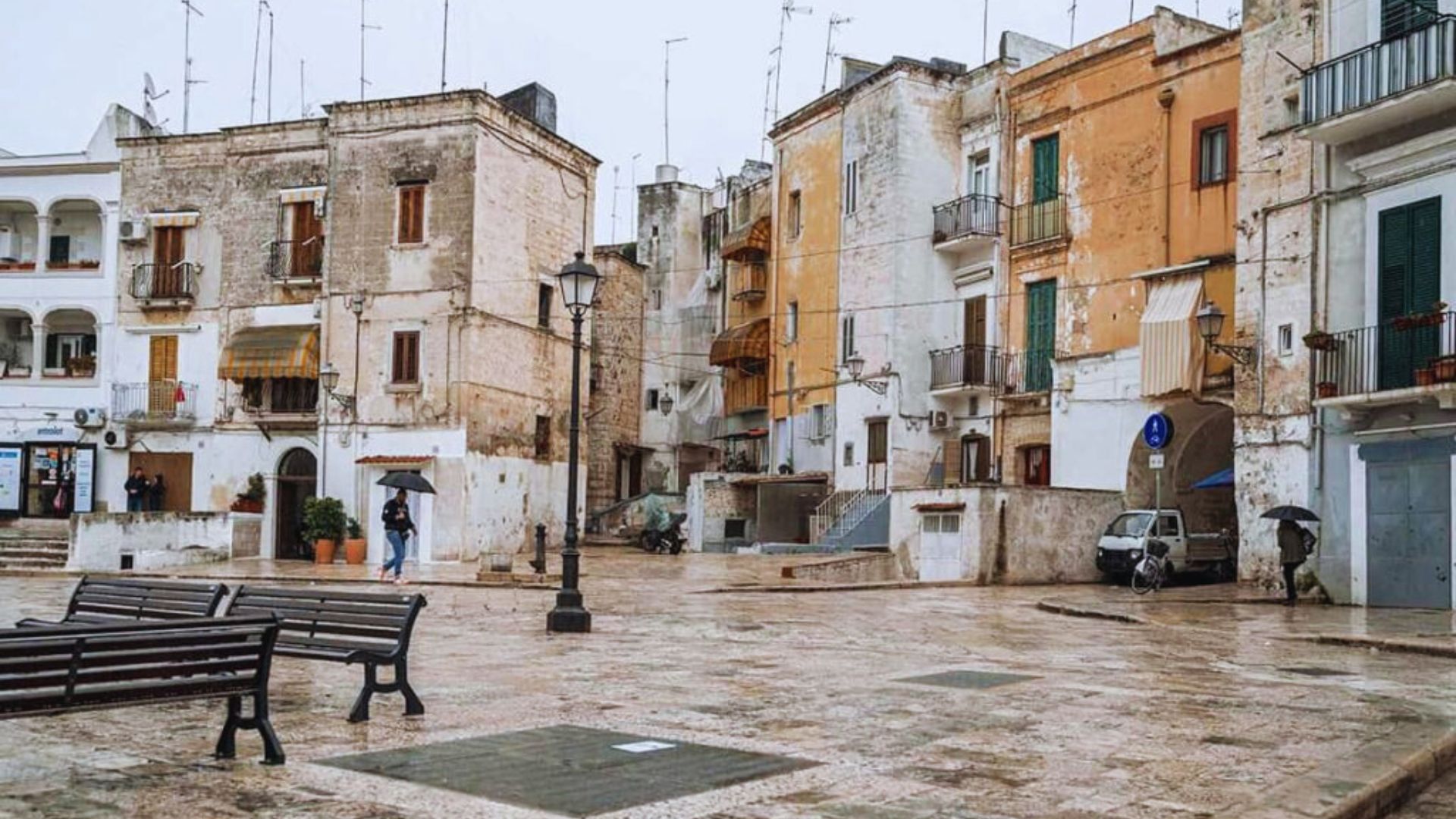 A rainy city square featuring benches and buildings, with puddles forming on the cobblestone pavement.