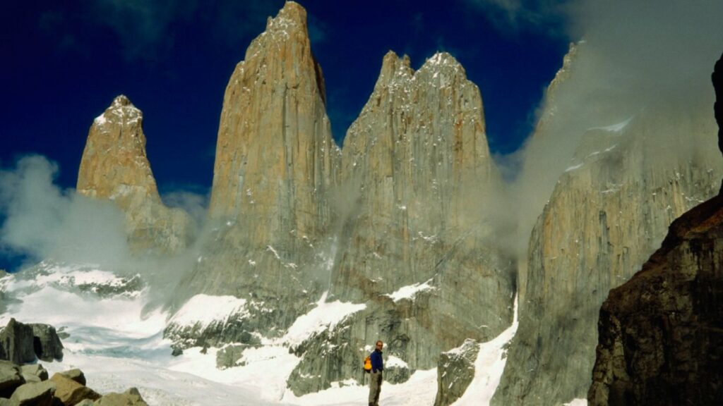 A man stands before towering mountains, showcasing the grandeur of nature in the background.