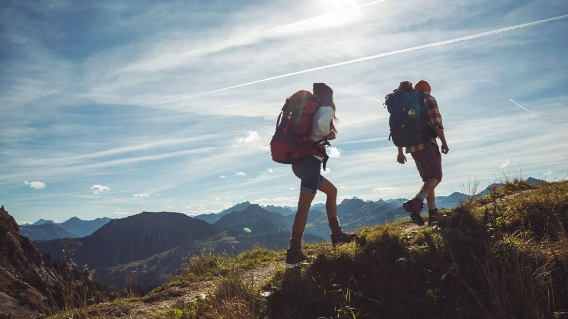 Two hikers with backpacks ascend a mountain trail, surrounded by rocky terrain and greenery.