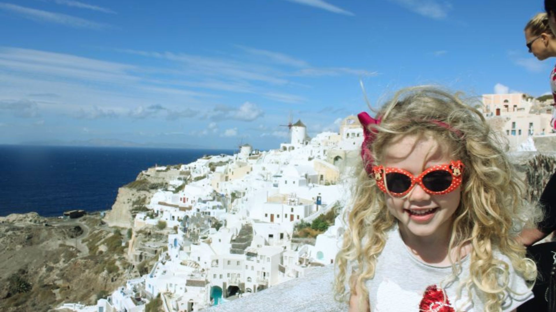 A young girl with curly blonde hair and red sunglasses smiles in the foreground. Behind her, the white buildings of Santorini, Greece, contrast with the deep blue sea and sky. The scene conveys joy and a sense of adventure.