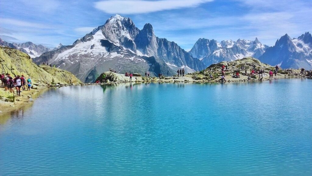People gather around a serene lake, with majestic mountains rising in the background under a clear blue sky.