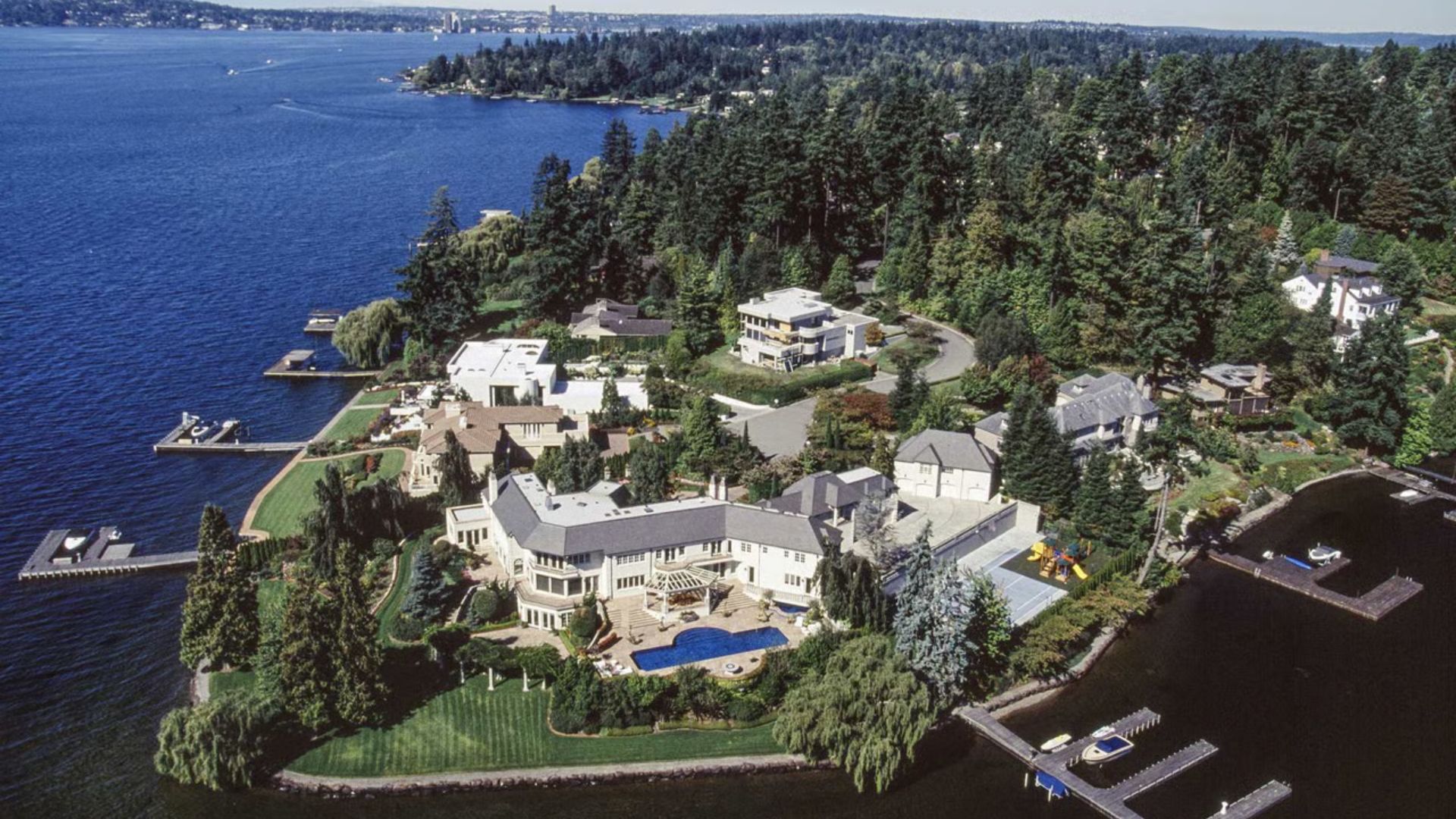 Aerial view of a large house situated on the water, surrounded by greenery and reflecting sunlight on the surface.