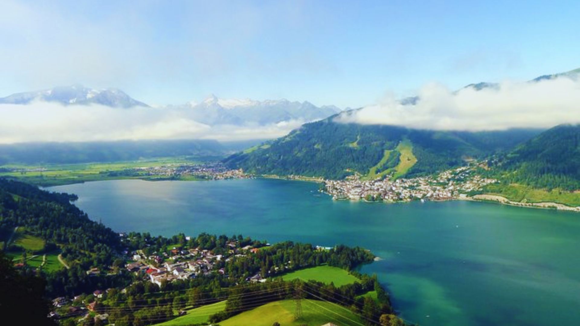 A panoramic view from a mountain peak, showcasing a serene lake surrounded by distant mountains under a clear sky.