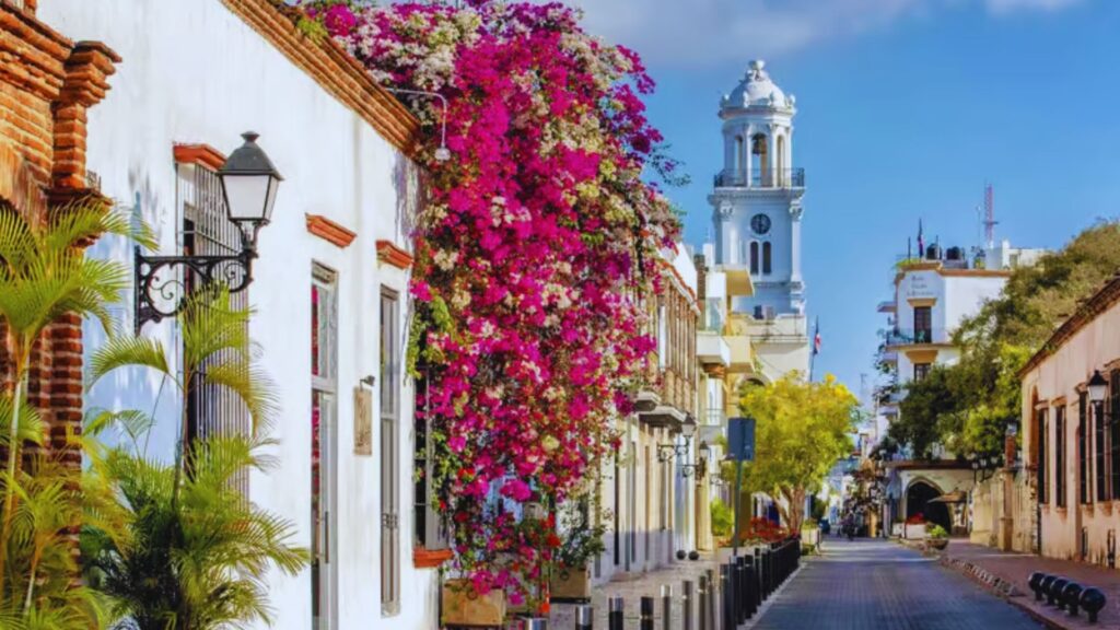 A picturesque street lined with colorful flowers, featuring a prominent church tower in the background.
