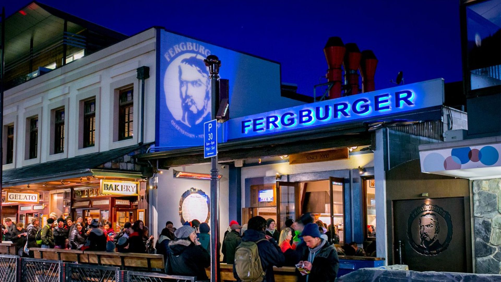 A group of people stands outside a restaurant at night, illuminated by warm lights and a bustling urban atmosphere.