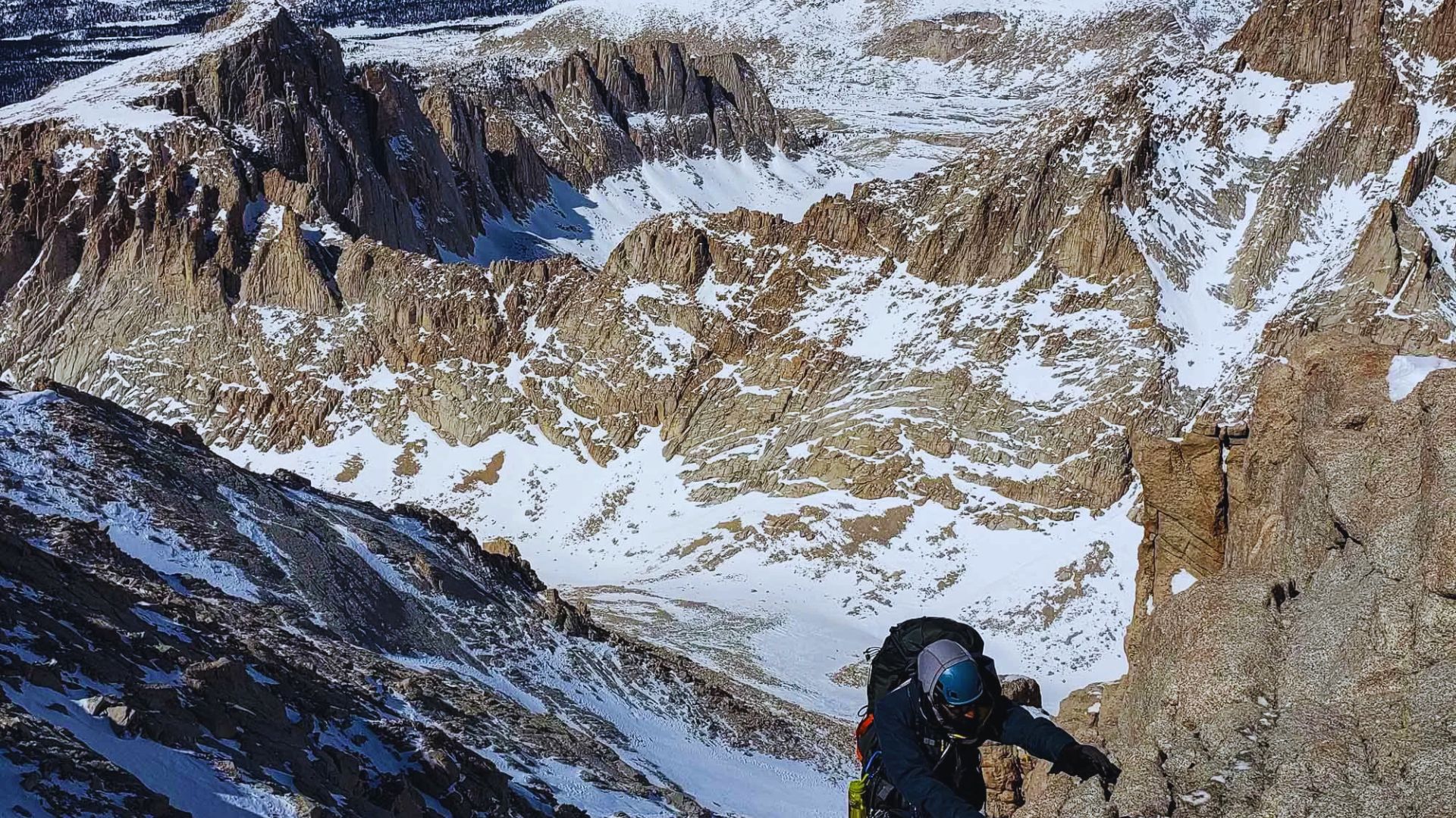 A climber ascends a steep mountain slope, with snow-covered peaks visible in the background.