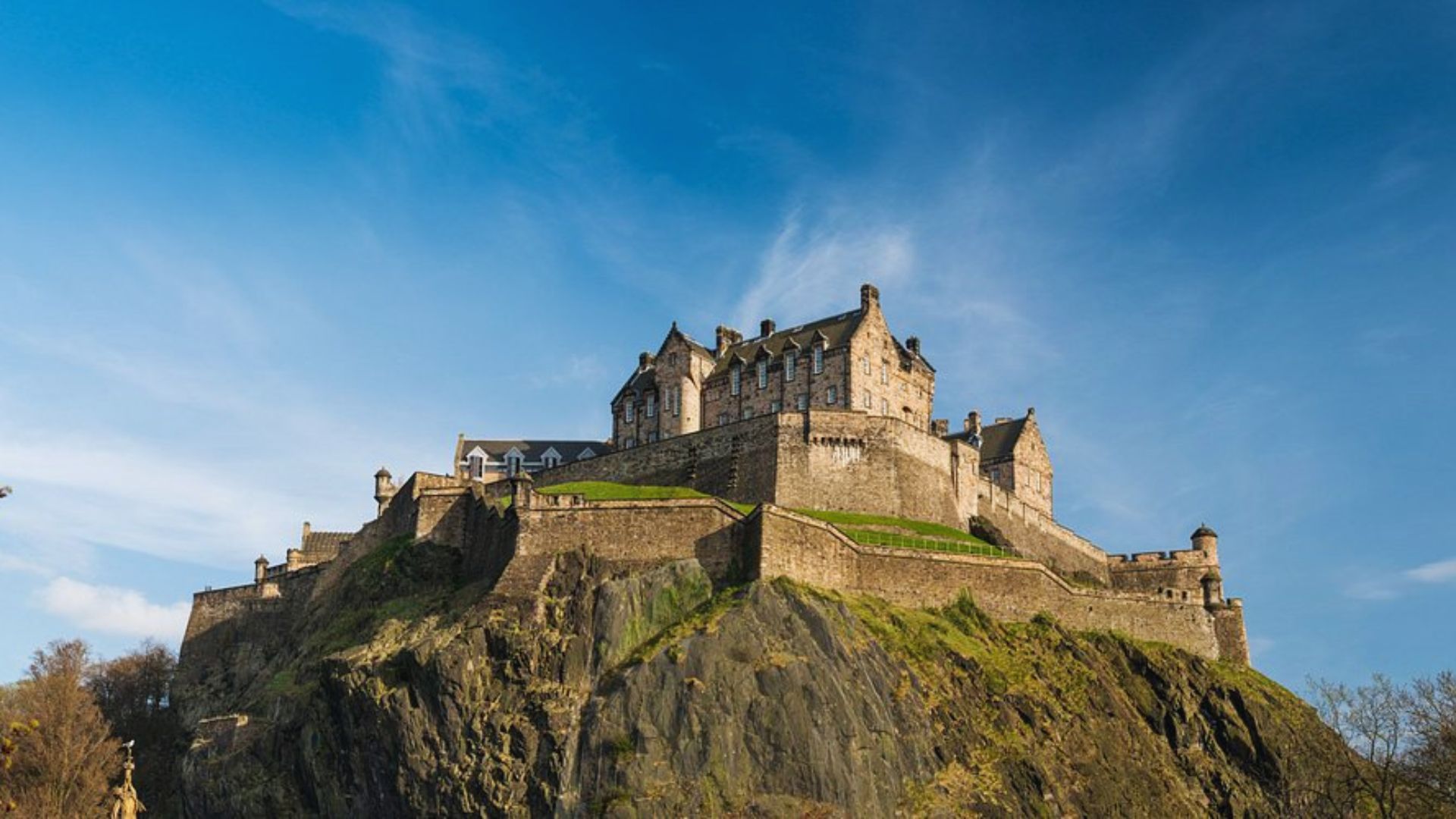 A majestic stone castle atop a rugged hill under a clear blue sky. The fortress exudes strength and history, framed by lush greenery at its base.