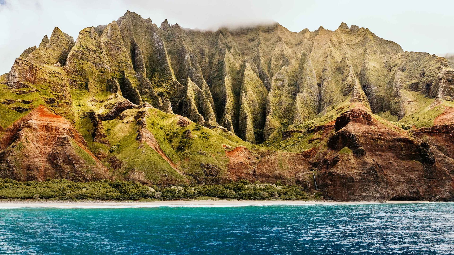 Dramatic coastal cliffs with jagged, green peaks rise above a sandy beach and turquoise ocean. The scene conveys tranquility and natural beauty.