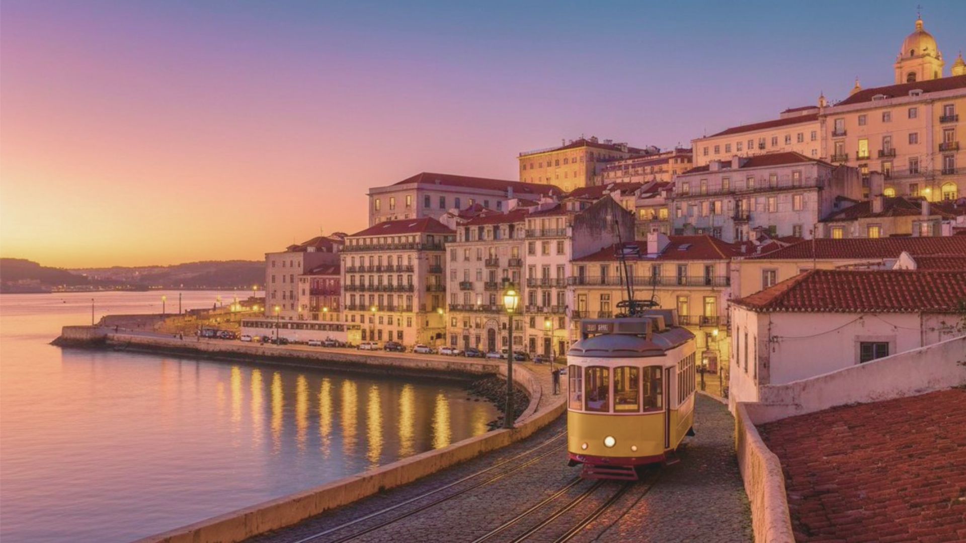 Sunset over Lisbon with a vintage tram on cobblestone streets lined with historic buildings. The sky glows pink and orange, reflecting on the river.