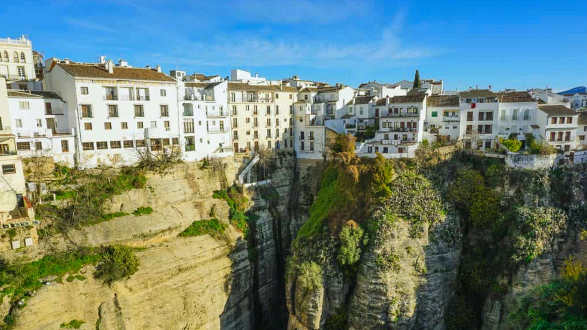 White buildings perched on rugged cliffs overlook a deep gorge, with lush greenery. A clear blue sky adds to the serene yet dramatic scene.