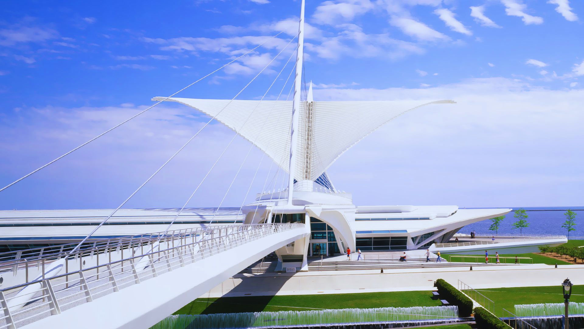 Futuristic white building with wing-like fins, set against a bright blue sky and lake. A long pedestrian bridge leads to the entrance. Modern and airy.