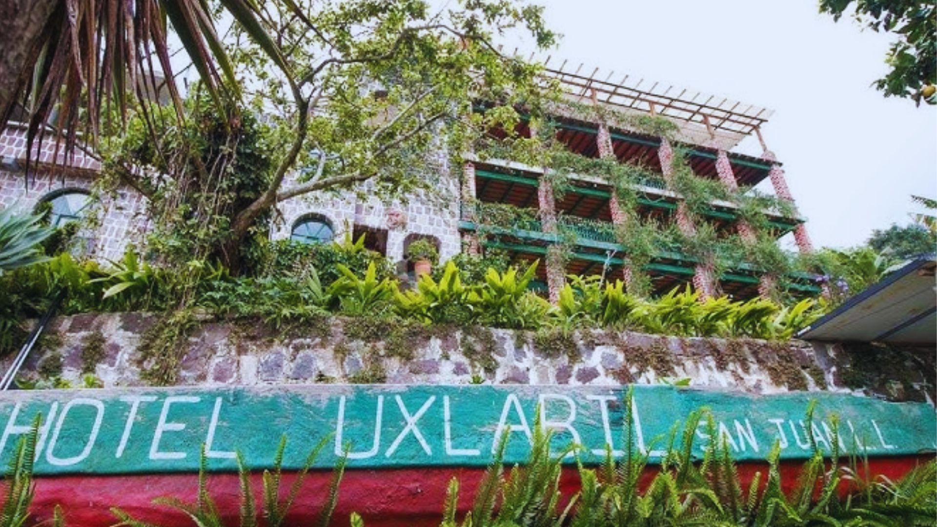 A rustic hotel exterior with green vines and plants covering stone walls and balconies, creating a lush, natural atmosphere. The sign reads "Hotel Uxlabil."