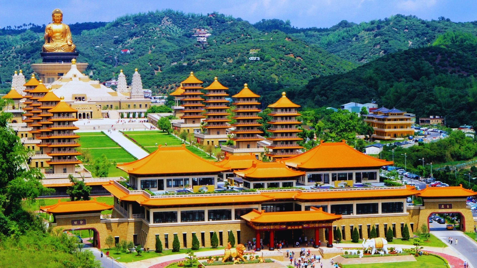 A sprawling Buddhist temple complex featuring multiple pagodas with orange roofs, lush green hills in the background, and a large golden Buddha statue.