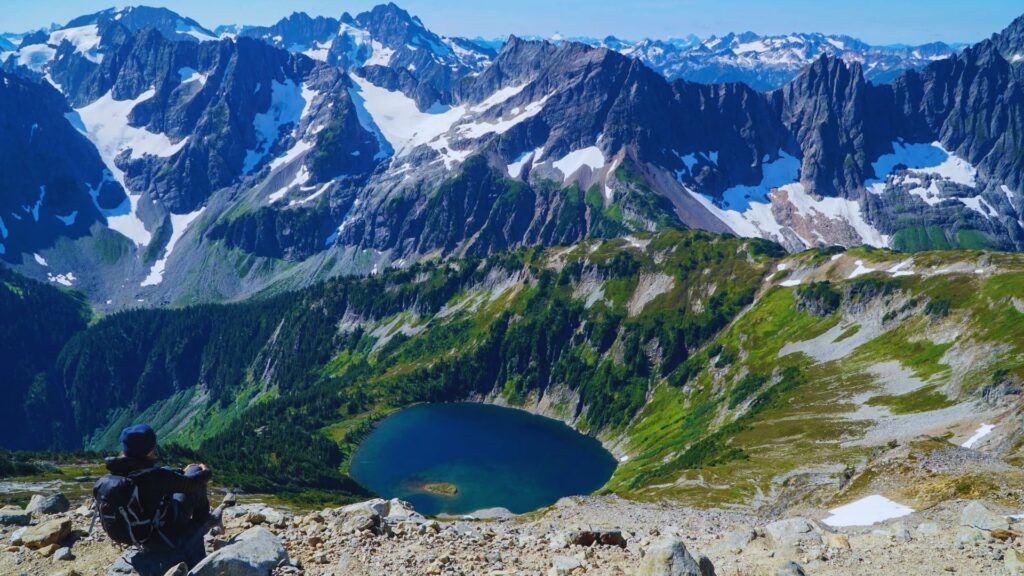 A person with a backpack sits on a rocky ledge overlooking a serene blue lake surrounded by lush greenery and towering snow-capped mountains under a blue sky.
