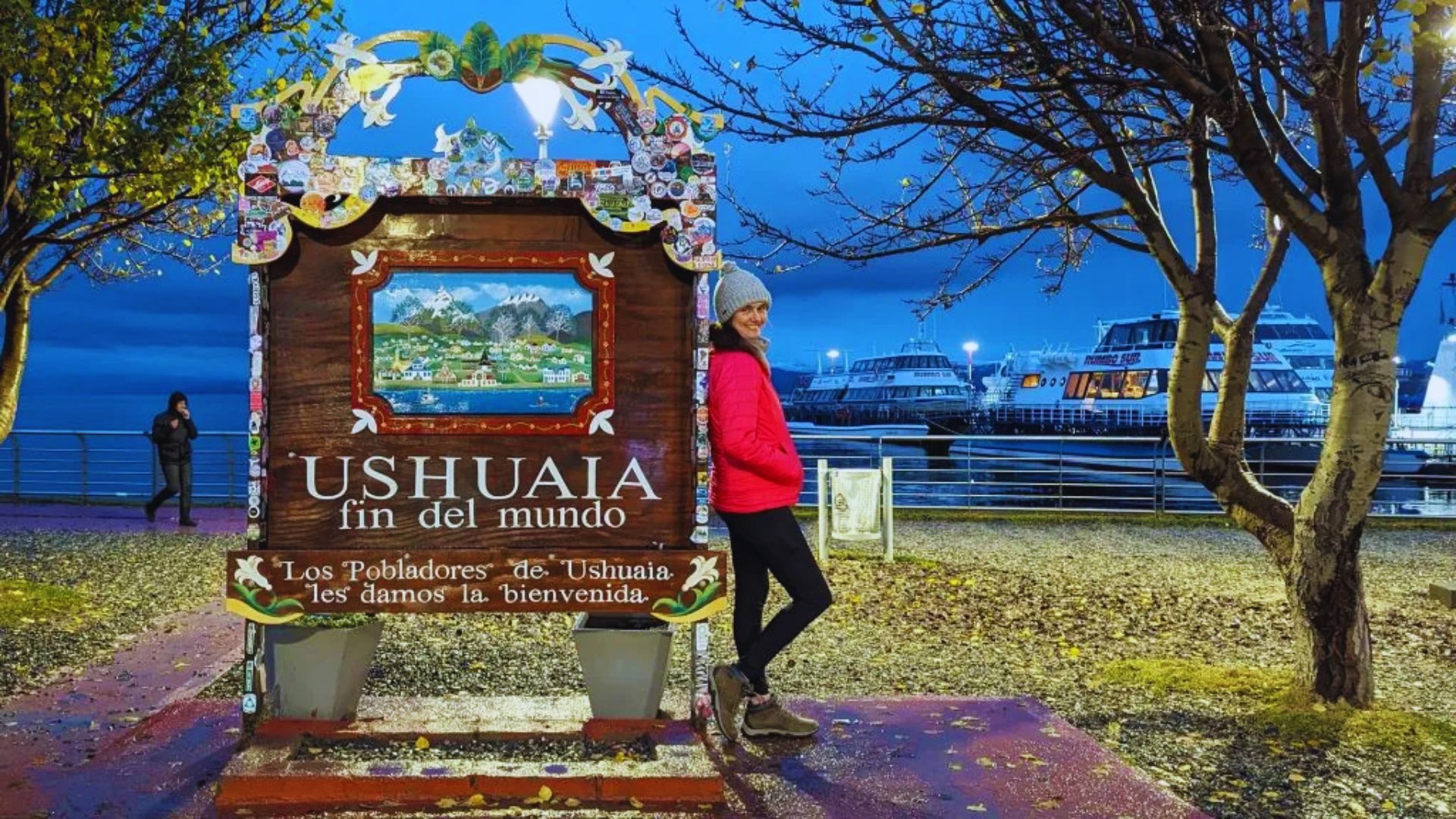A woman in a pink jacket and gray hat stands smiling beside the Ushuaia "fin del mundo" sign. Behind her are docked boats and leafless trees at dusk.