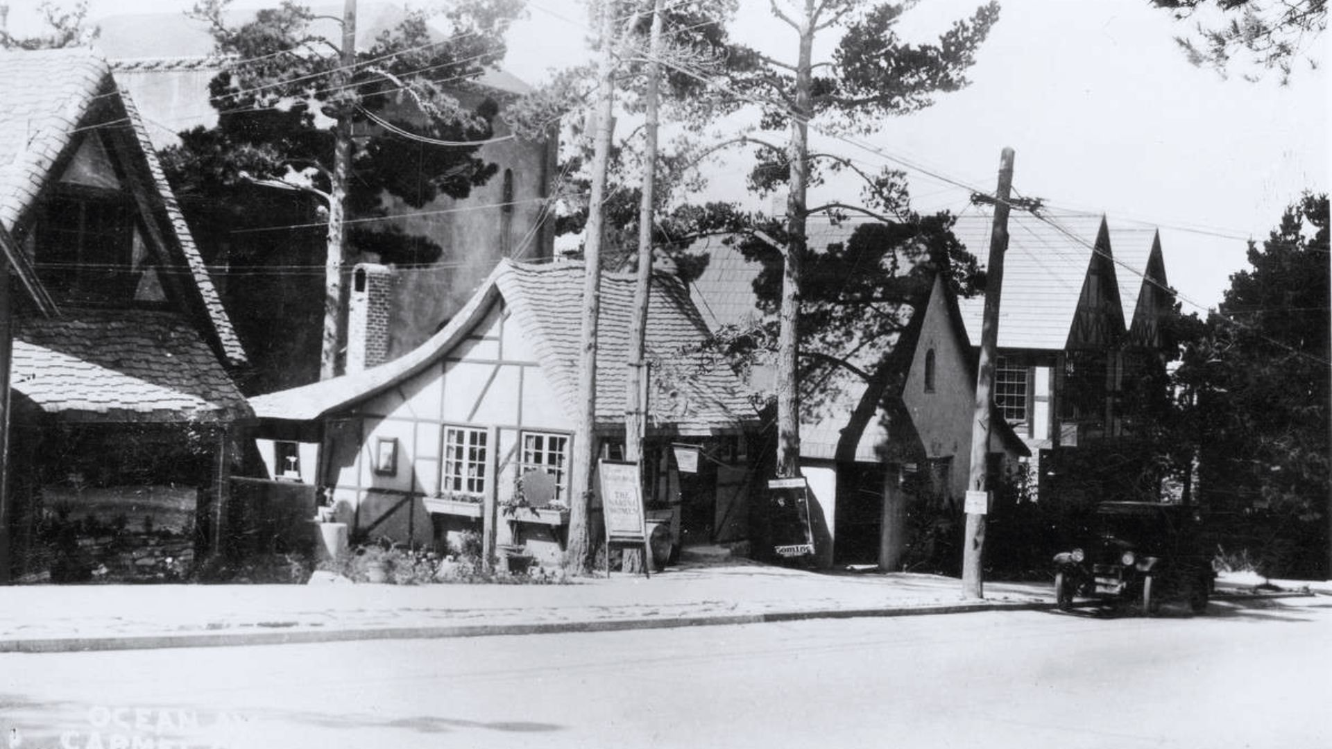 Historic black-and-white photo of quaint, European-style cottages lined along a street, nestled among tall pine trees; an old car is parked nearby.