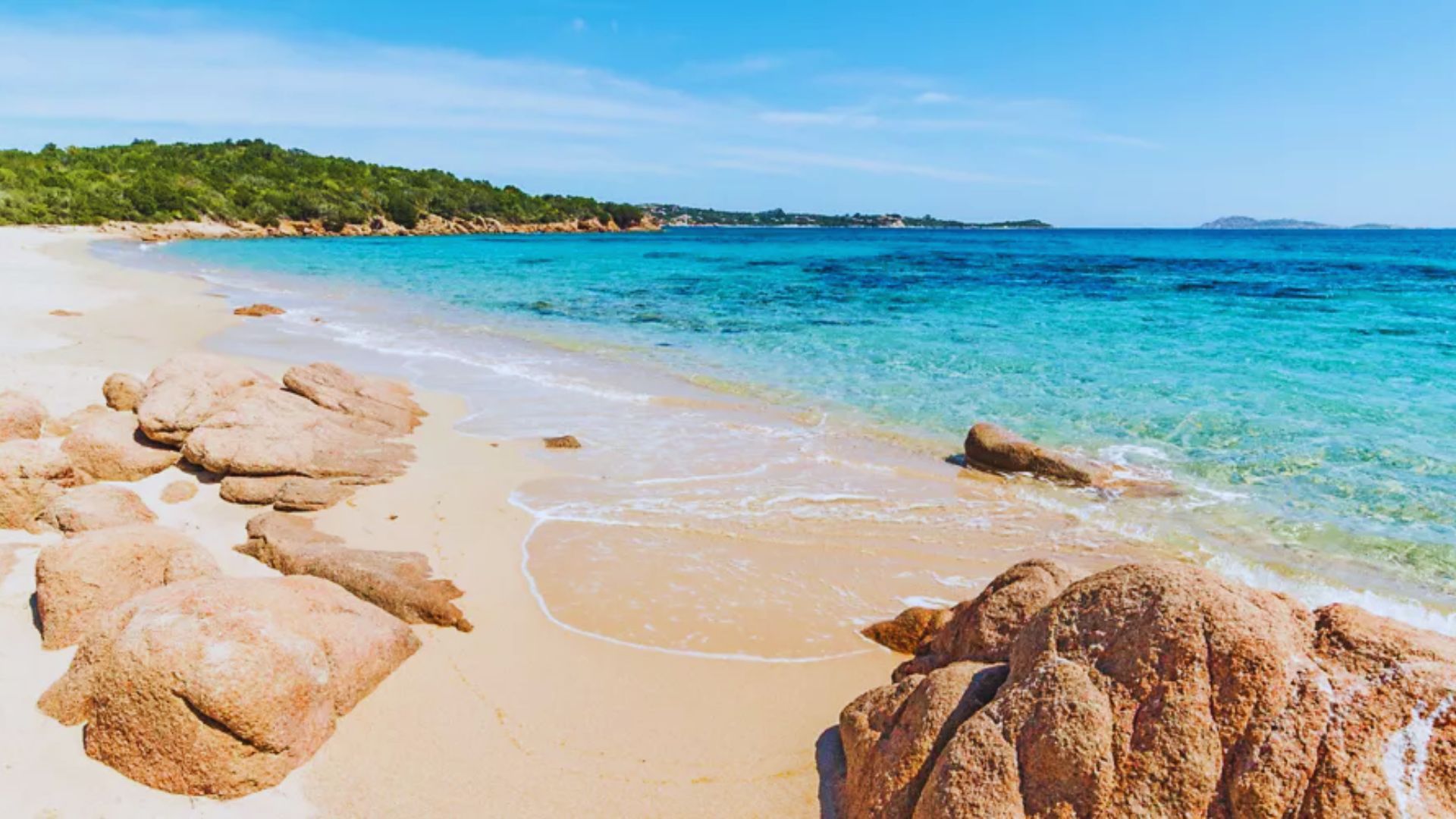 Sunny beach scene with clear blue water, gentle waves, and golden sand. Smooth rocks in the foreground and lush greenery on the distant shoreline. Peaceful ambiance.