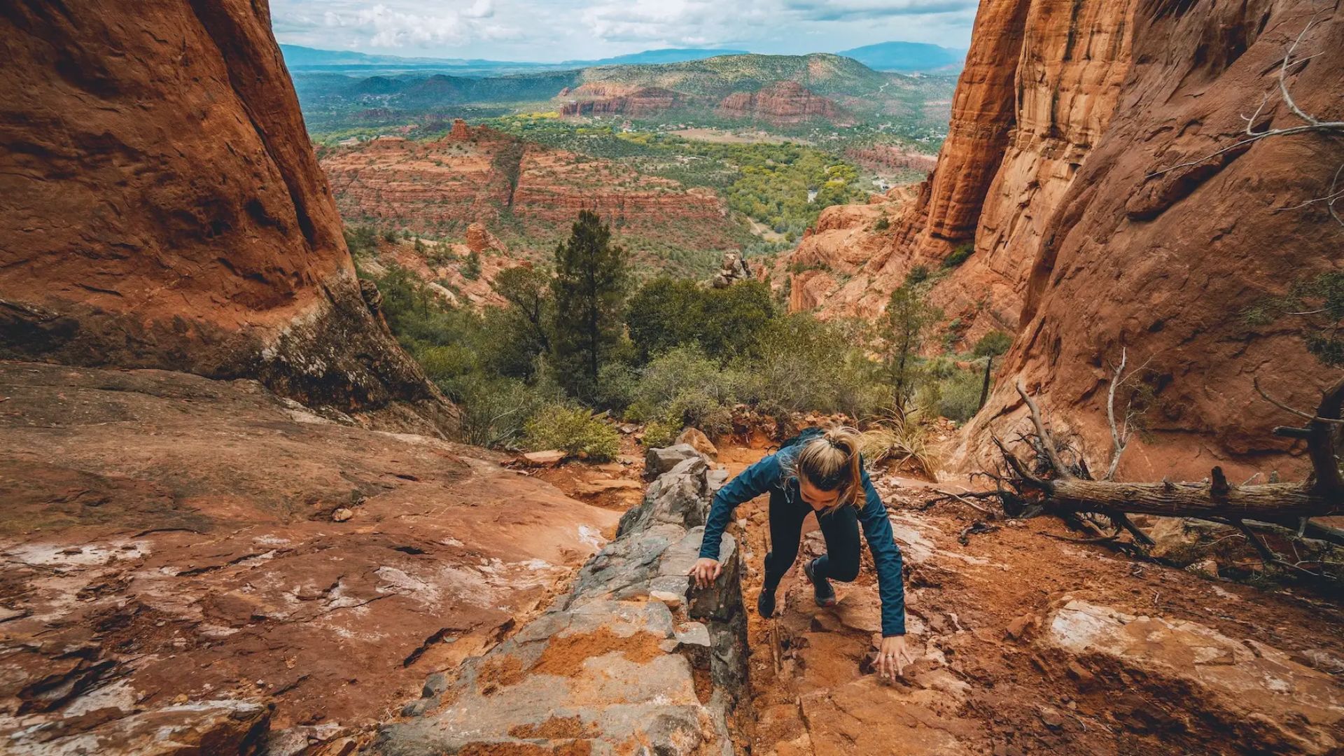 A person climbs a steep, rocky path between red canyon walls under a cloudy sky. Greenery and distant mountains form a stunning backdrop, evoking adventure.
