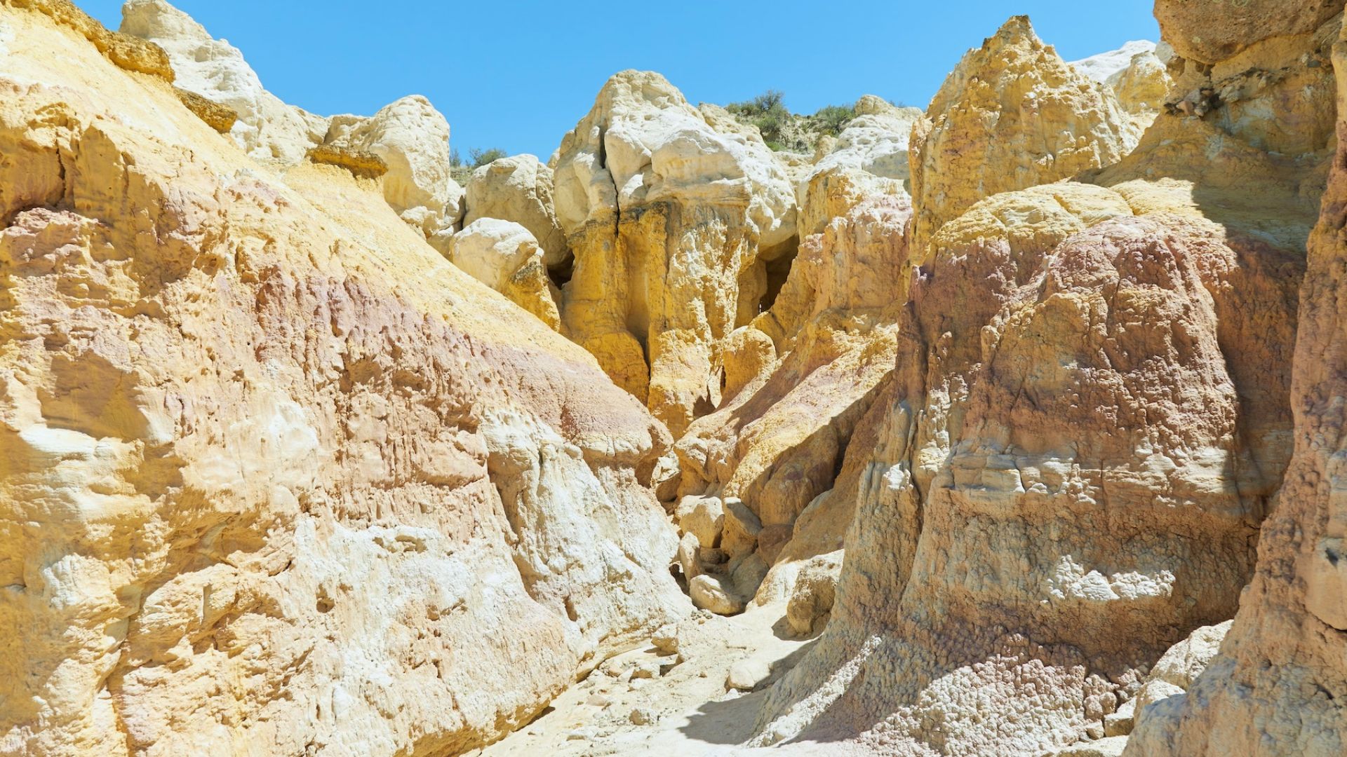 Colorful, rugged rock formations under a clear blue sky. The rocks display shades of yellow, pink, and orange, creating a striking natural landscape.
