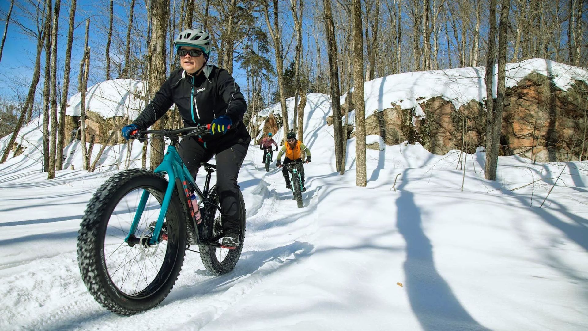 A group of cyclists ride fat tire bikes on a snowy trail through a forest. Sunlight filters through the trees, creating a joyful and adventurous scene.