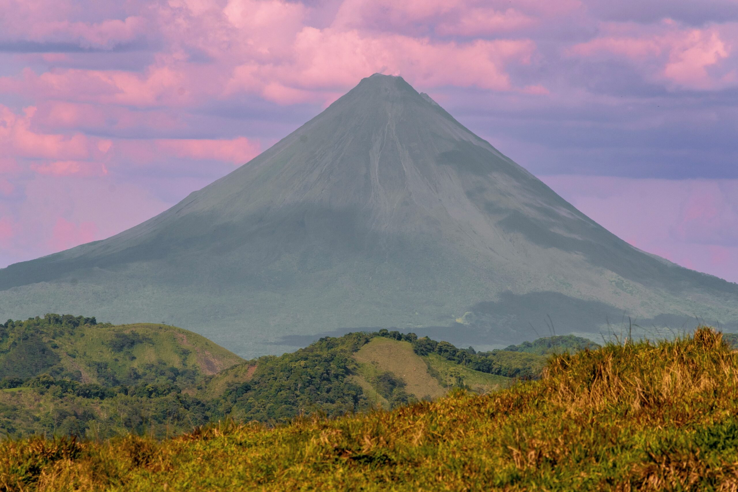 A majestic volcano rises against a vibrant pink and purple sky. Lush green hills in the foreground create a serene and dramatic landscape.