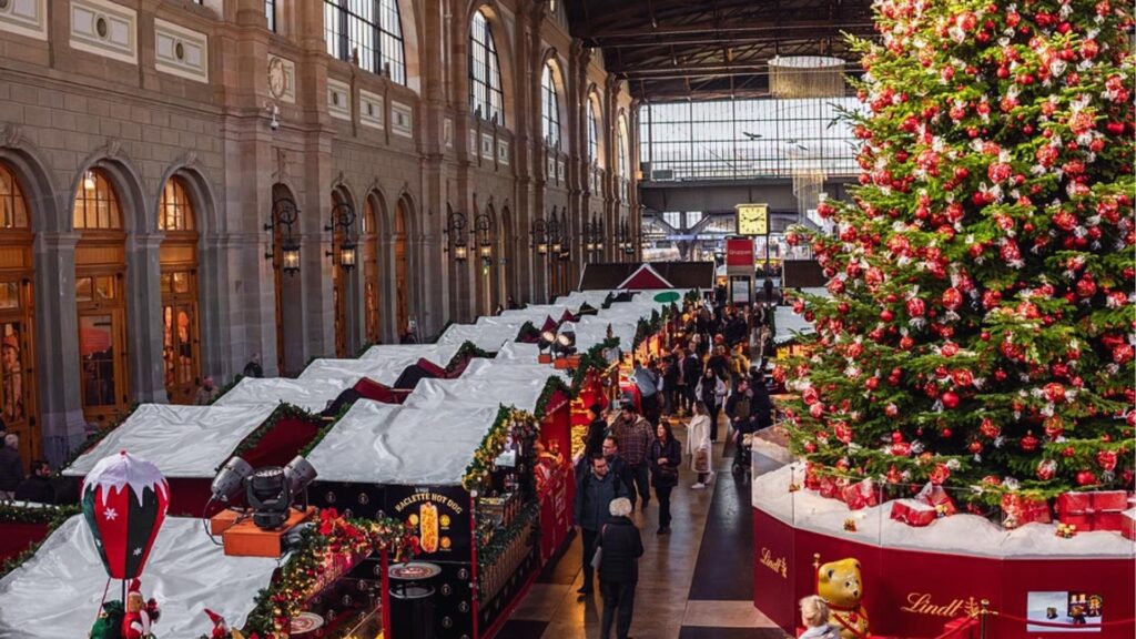 A bustling Christmas market in a train station featuring a large, decorated Christmas tree in the center.