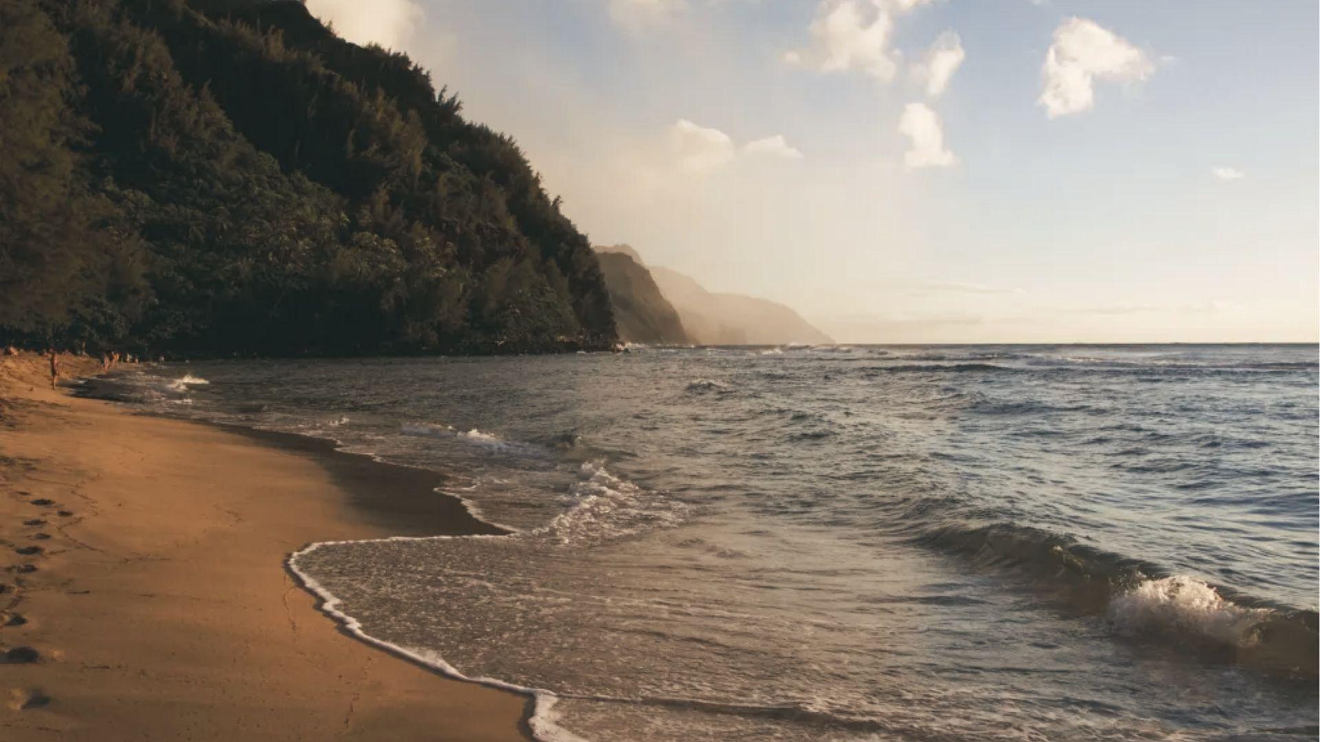 Golden sand beach with gentle waves lapping the shore, backed by steep green cliffs.
