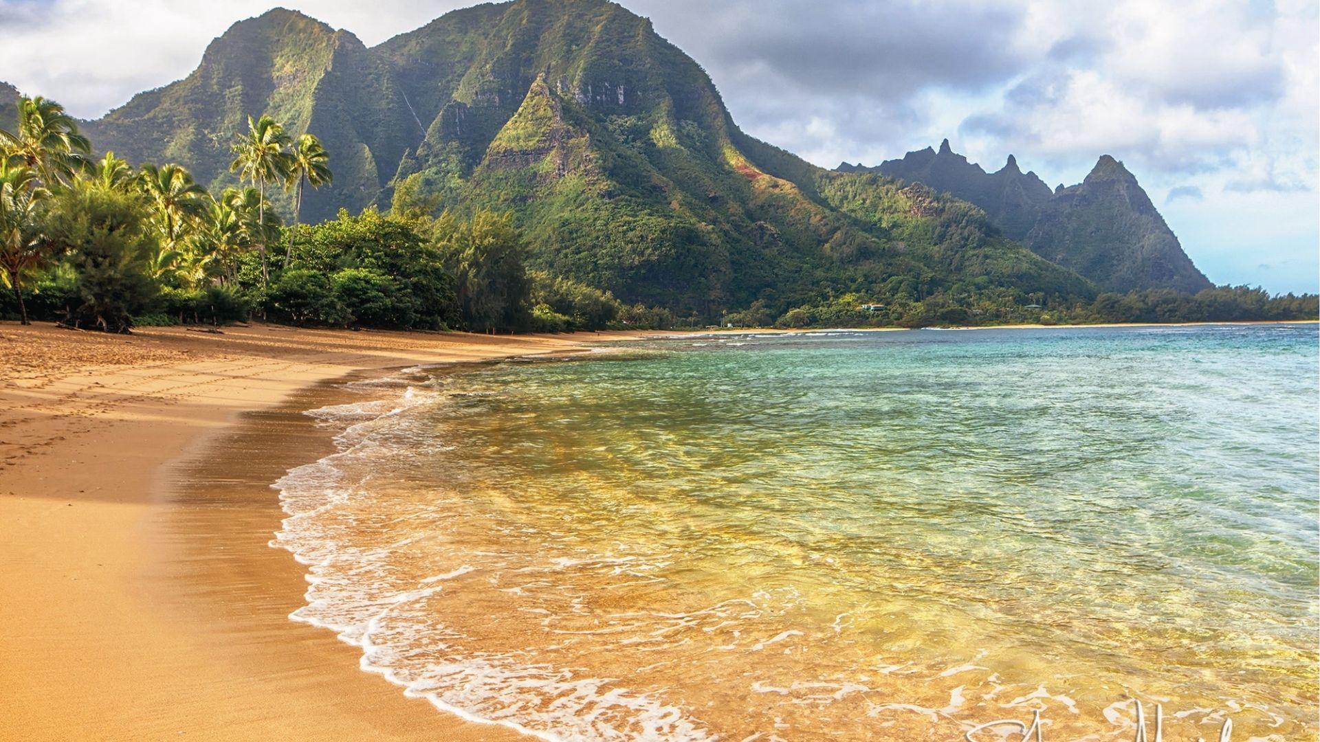 Sandy beach curves along clear water with lush, jagged mountains rising behind it.