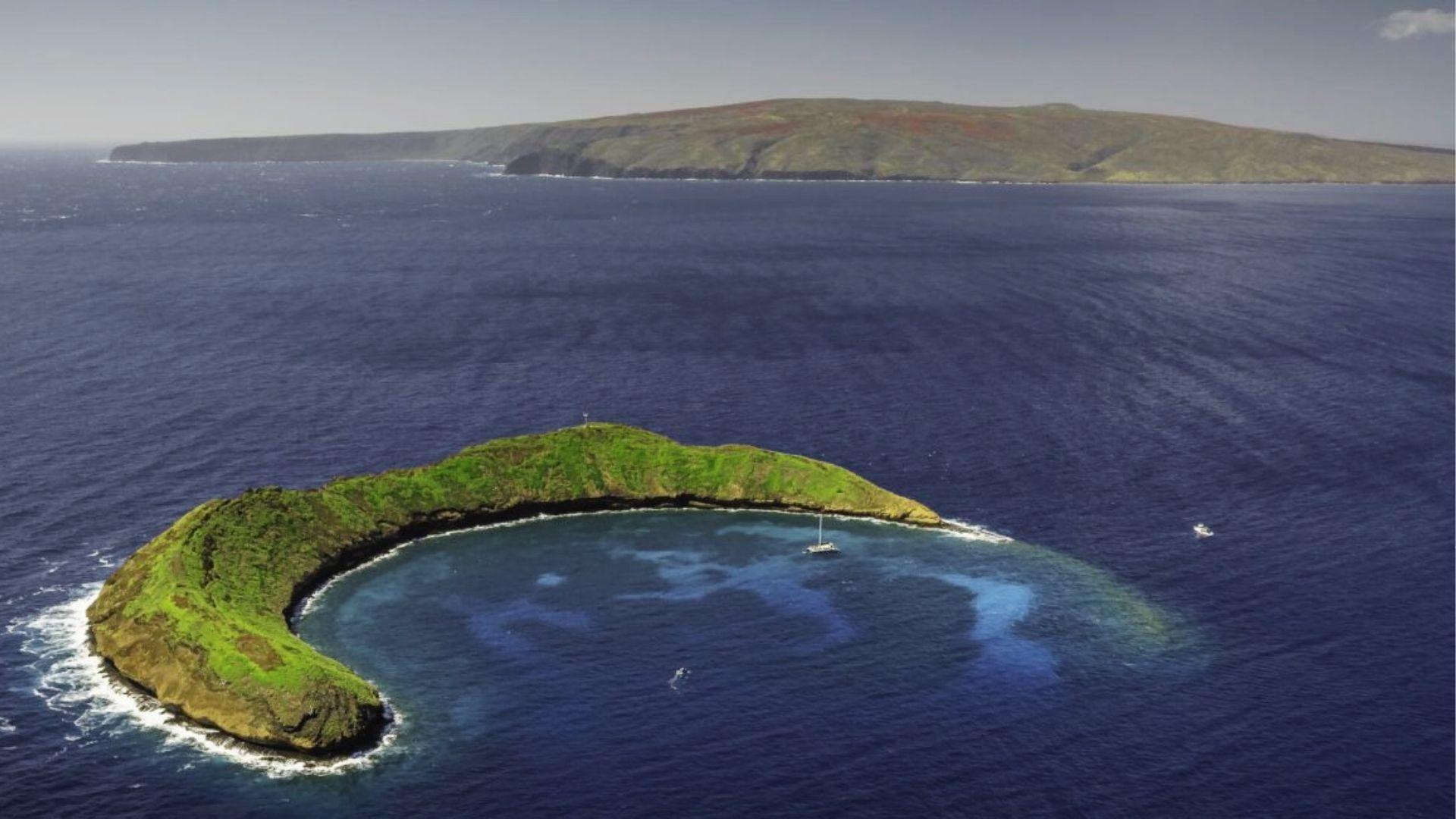 Aerial of a crescent-shaped green islet surrounded by deep blue ocean and a sailboat.